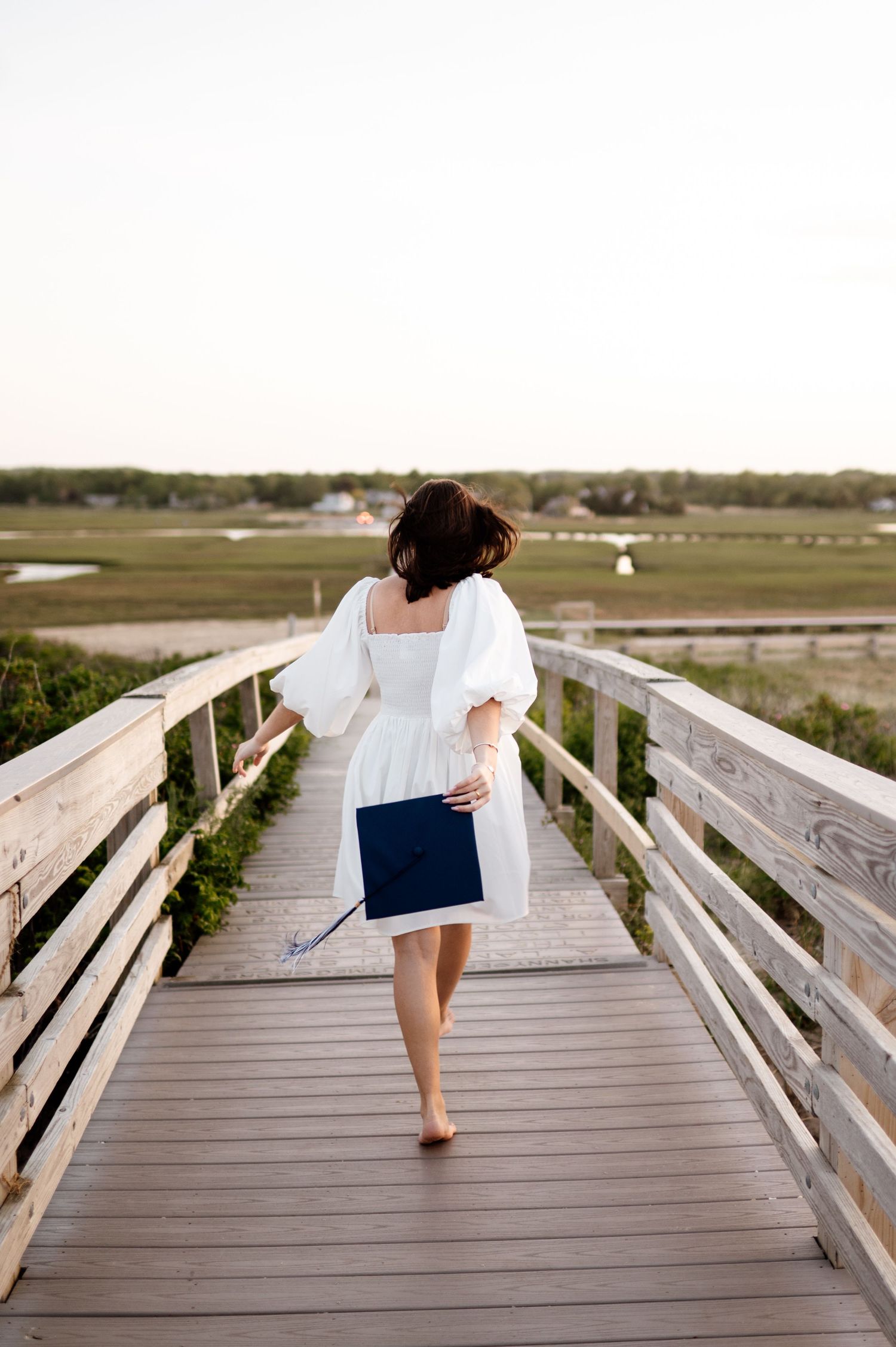 A person in a white dress walks barefoot on a wooden boardwalk overlooking a coastal marsh.