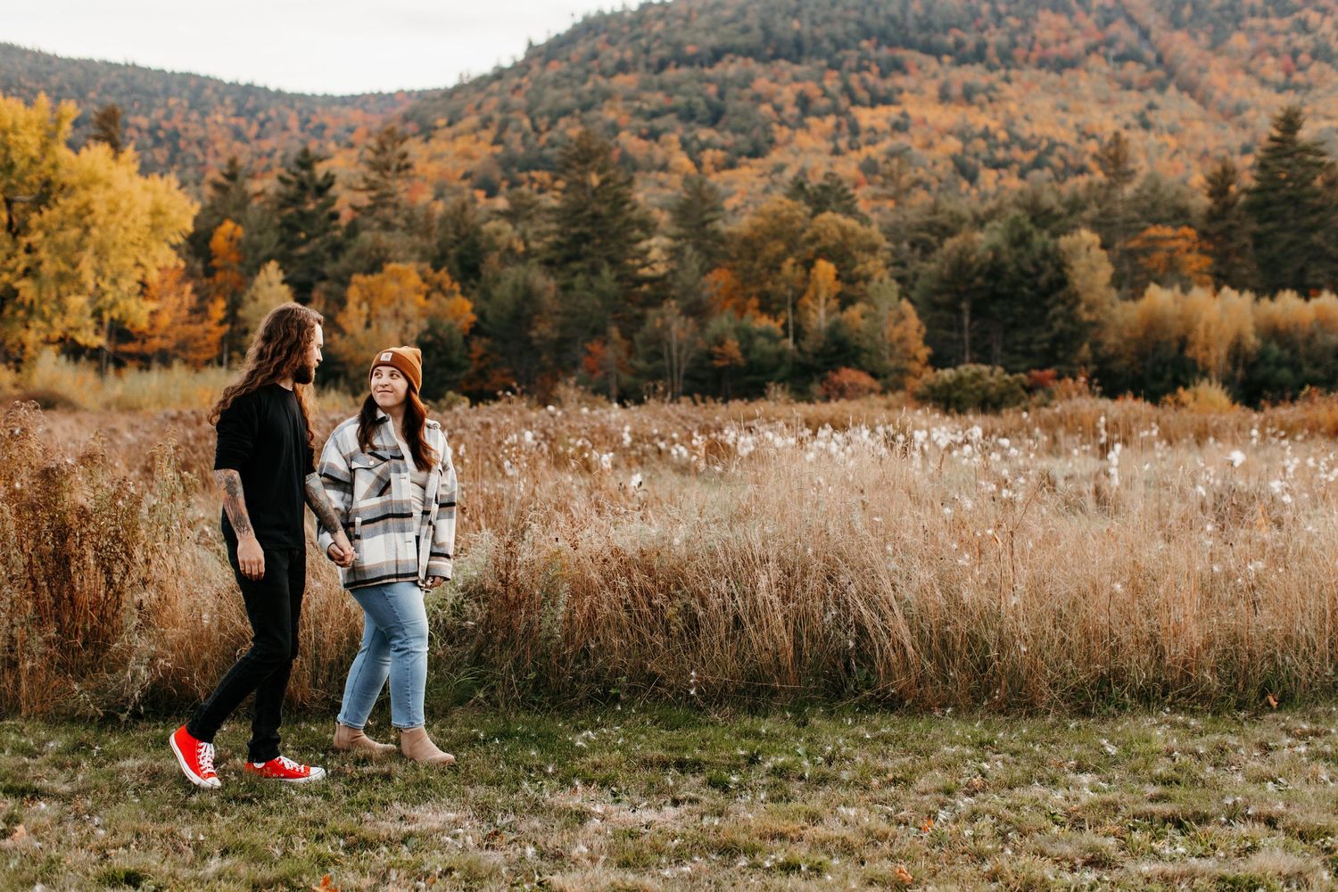 A couple walks through a scenic autumn meadow with golden grass and mountains in the background during sunset.