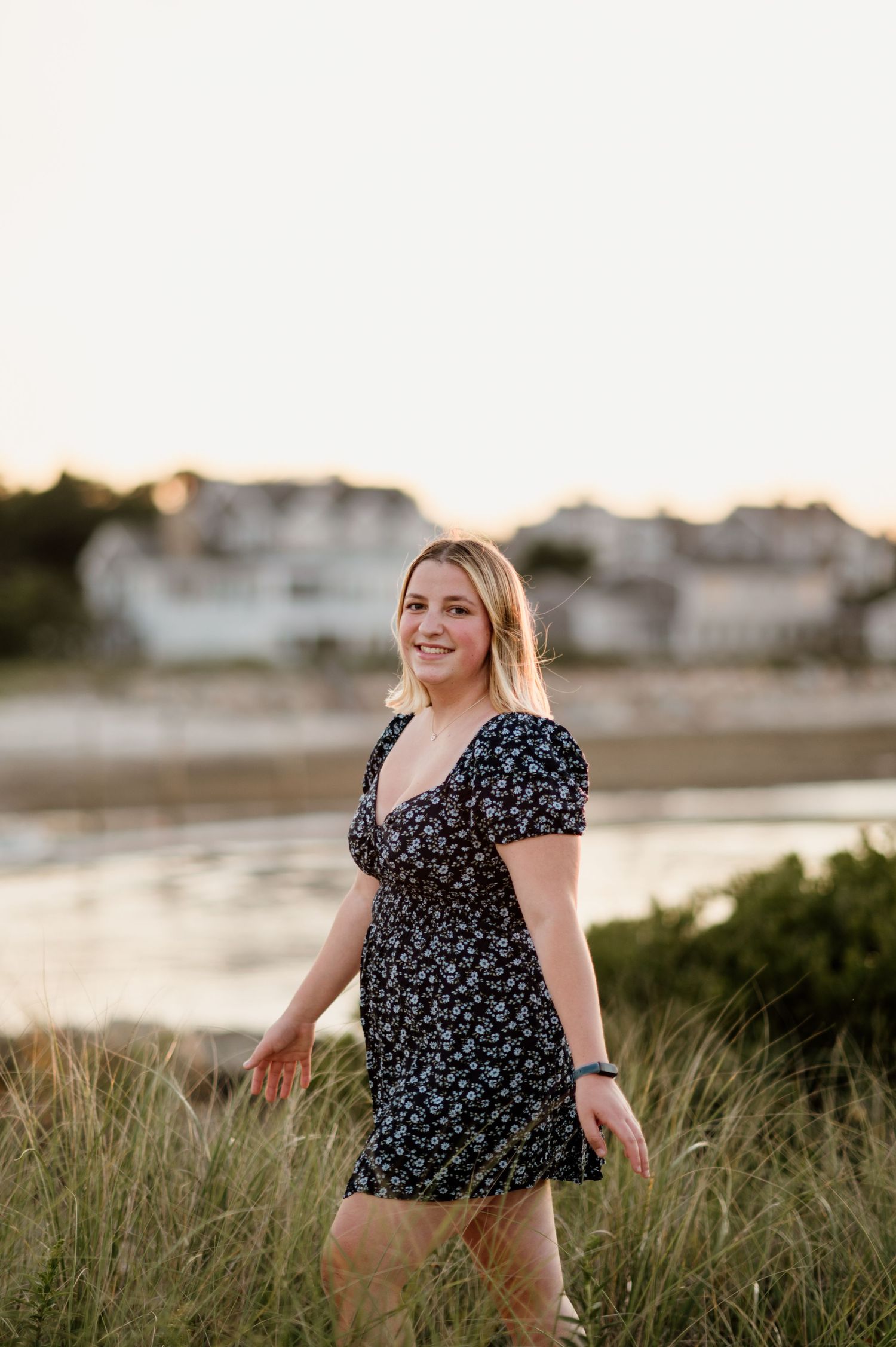 A person in a floral dress stands in coastal beach grass during golden hour sunset.