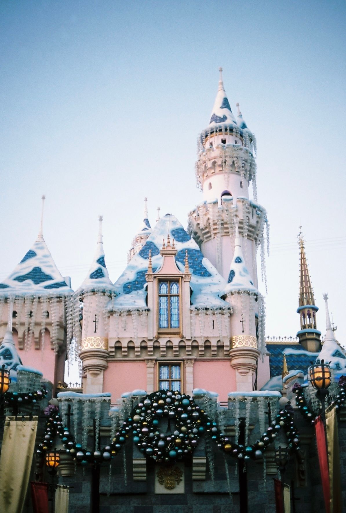 Panoramic view of Disneyland's Sleeping Beauty Castle with blue spires and pink walls against a light blue sky.