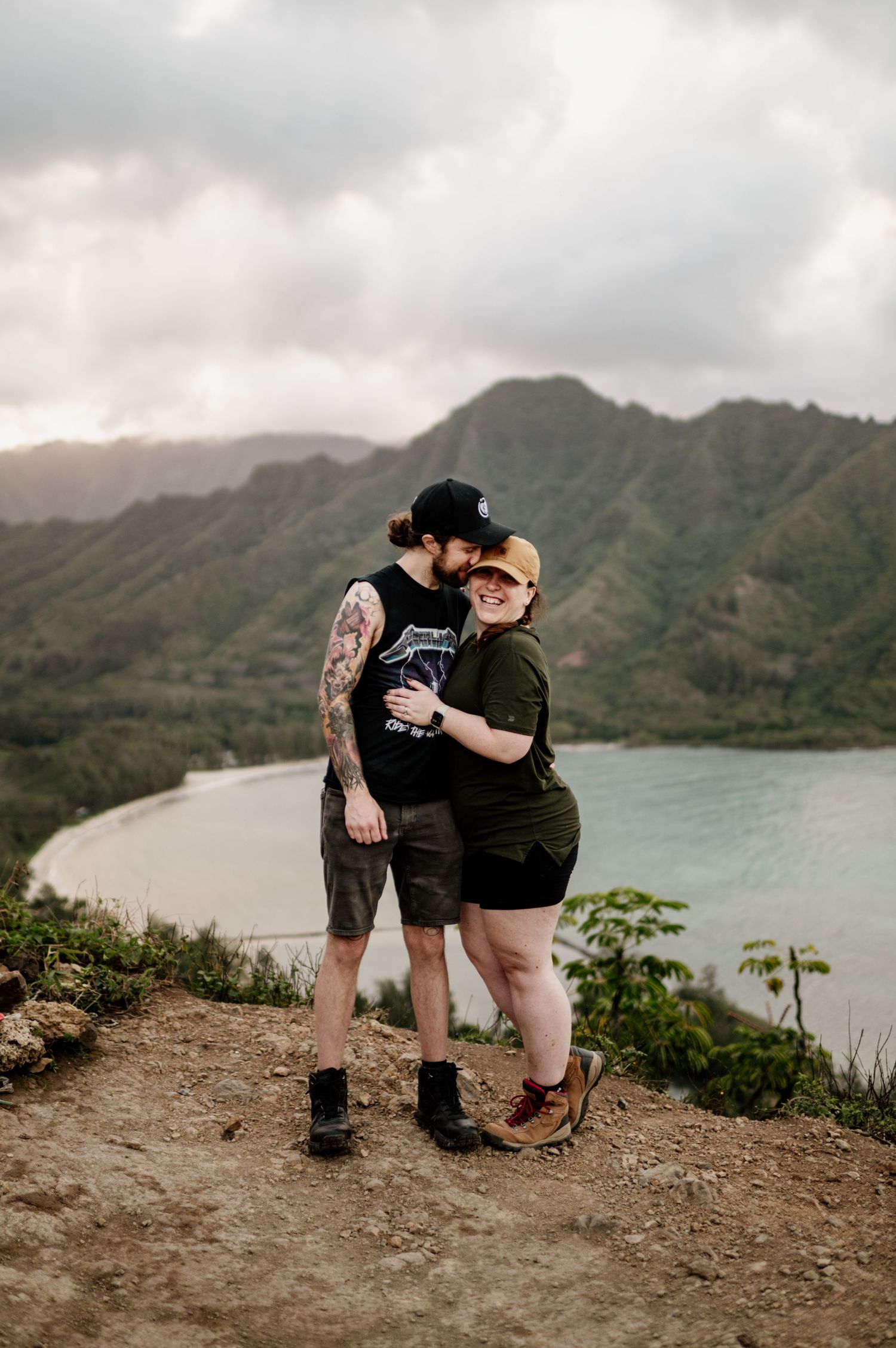 Two people embrace on a scenic mountain overlook with a turquoise lake and green mountains in the background.