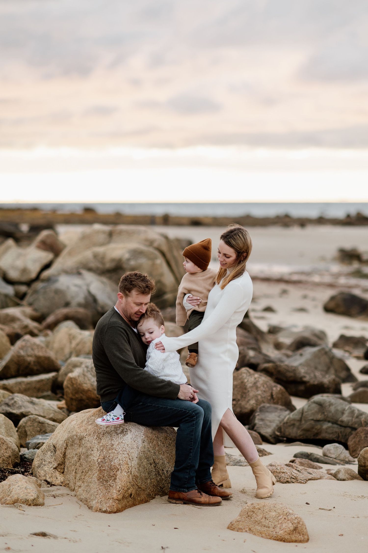 A romantic couple sits together on coastal rocks at sunset during a beach photoshoot.