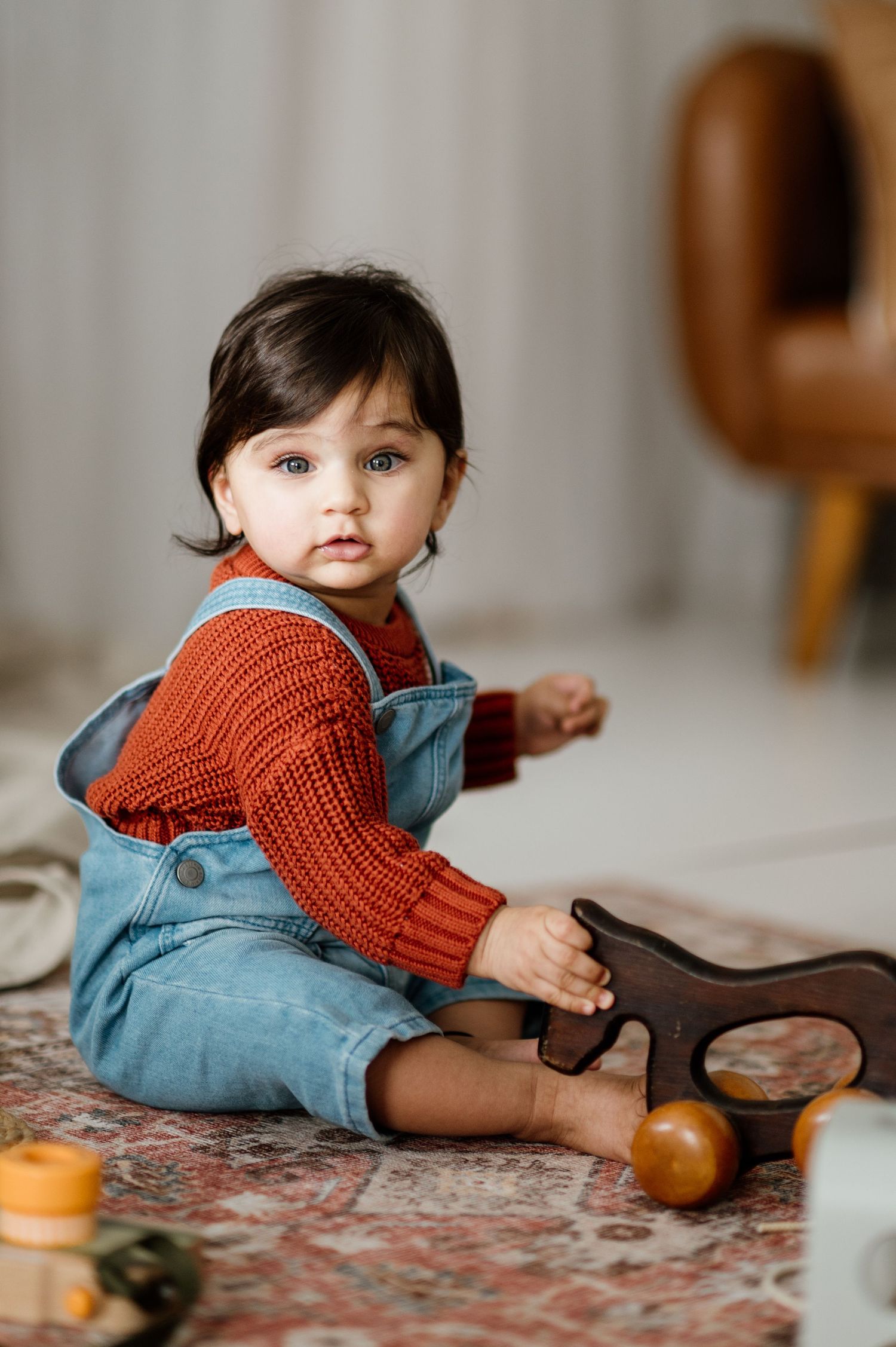 Baby wearing denim overalls and red sweater playing with toys on patterned carpet.