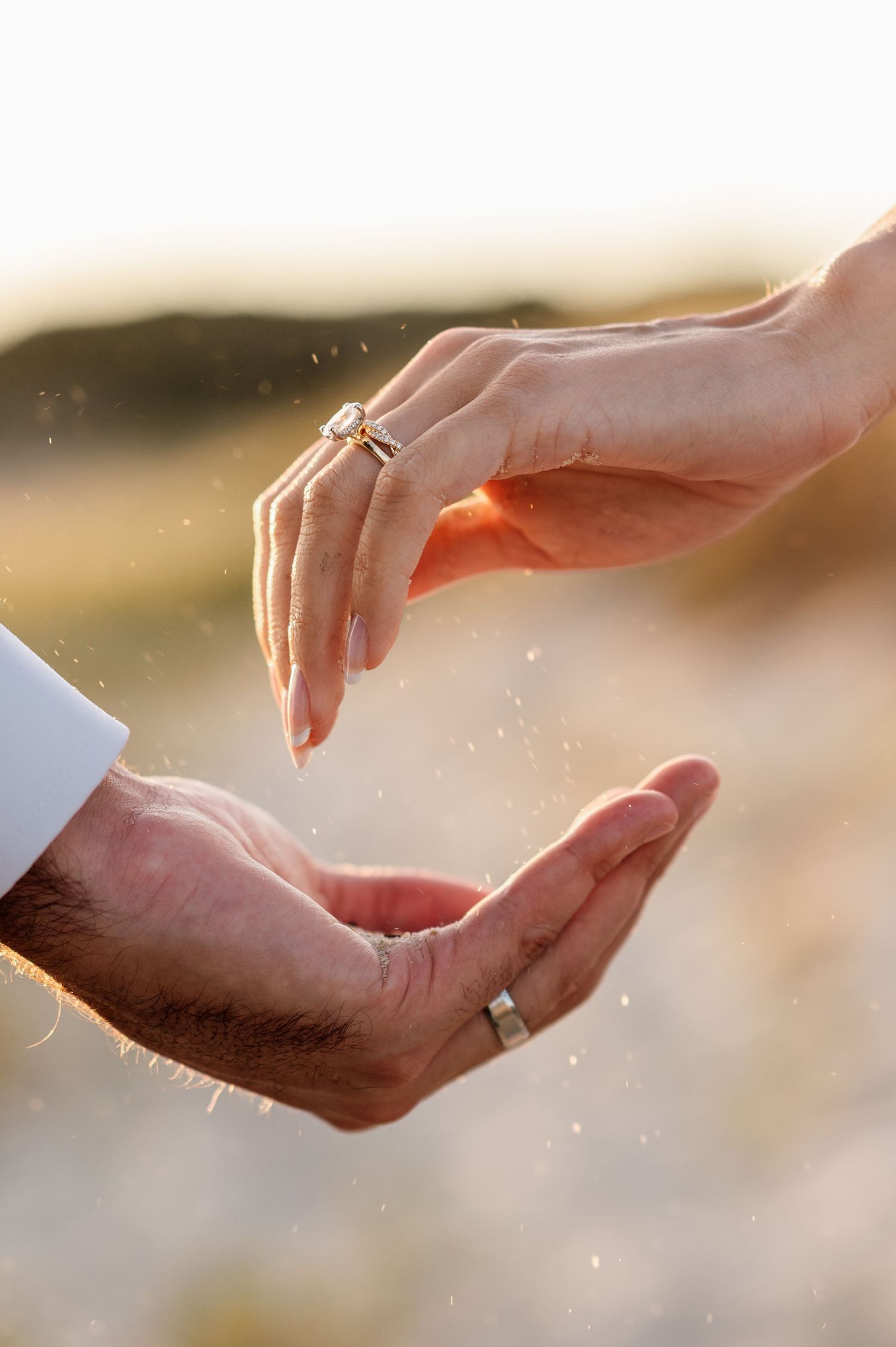 Close-up of couple's hands with wedding rings, sand falling between them in golden sunset light.