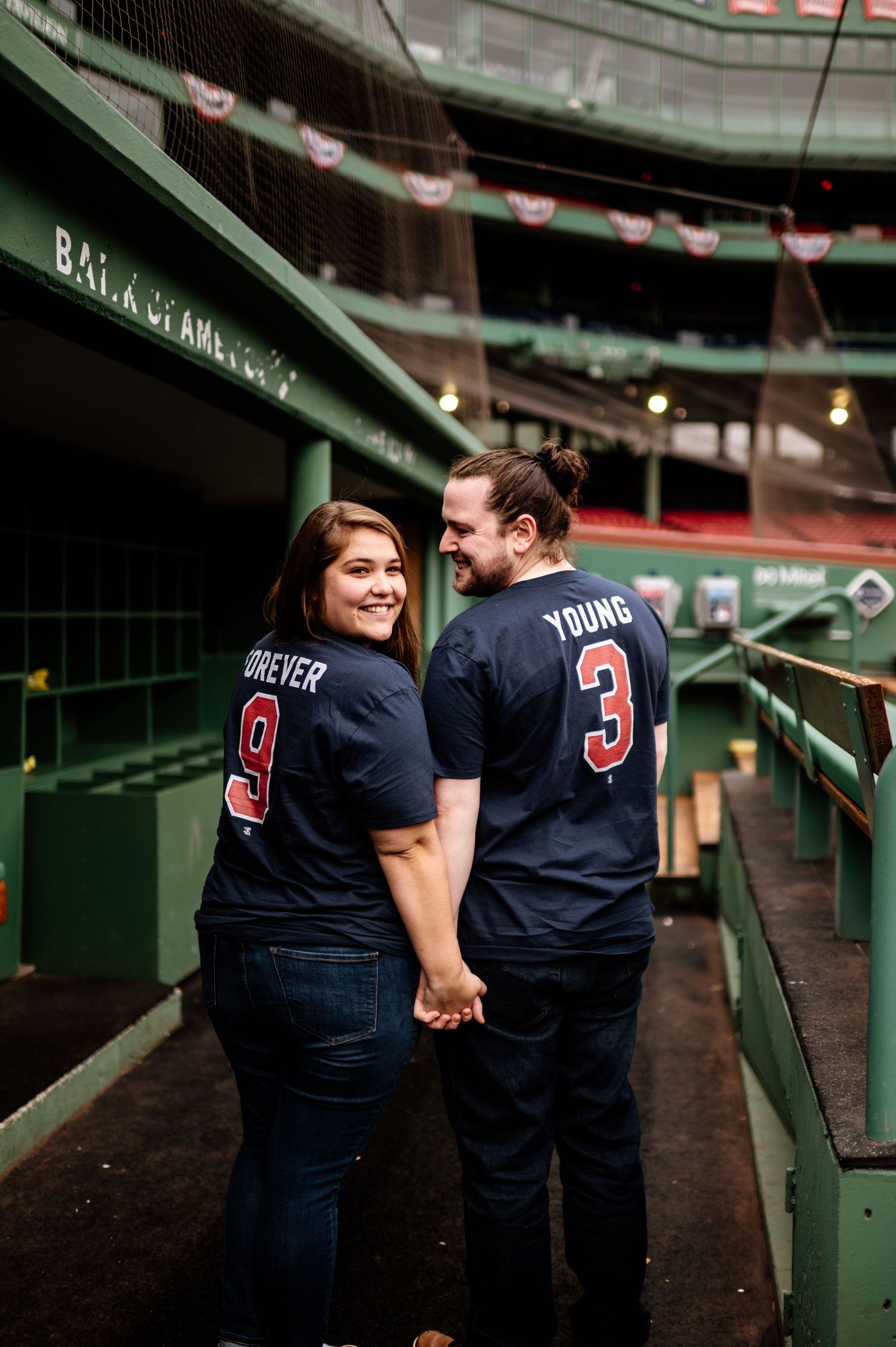 A couple wearing matching baseball jerseys hold hands while standing in Fenway Park stadium.