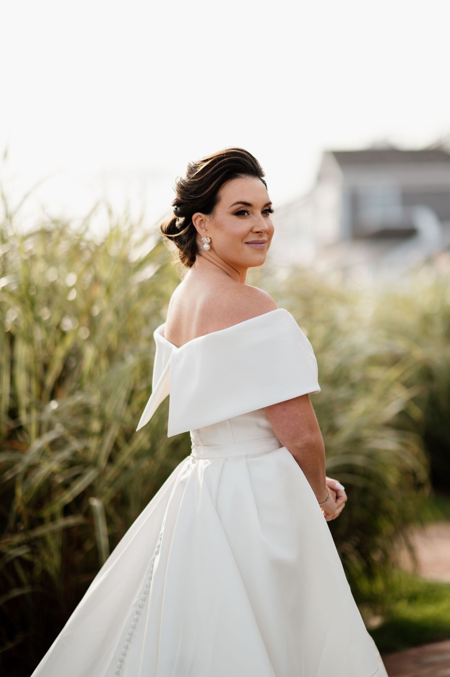 A bride in an elegant off-shoulder white wedding gown poses against a backdrop of tall grass at golden hour.
