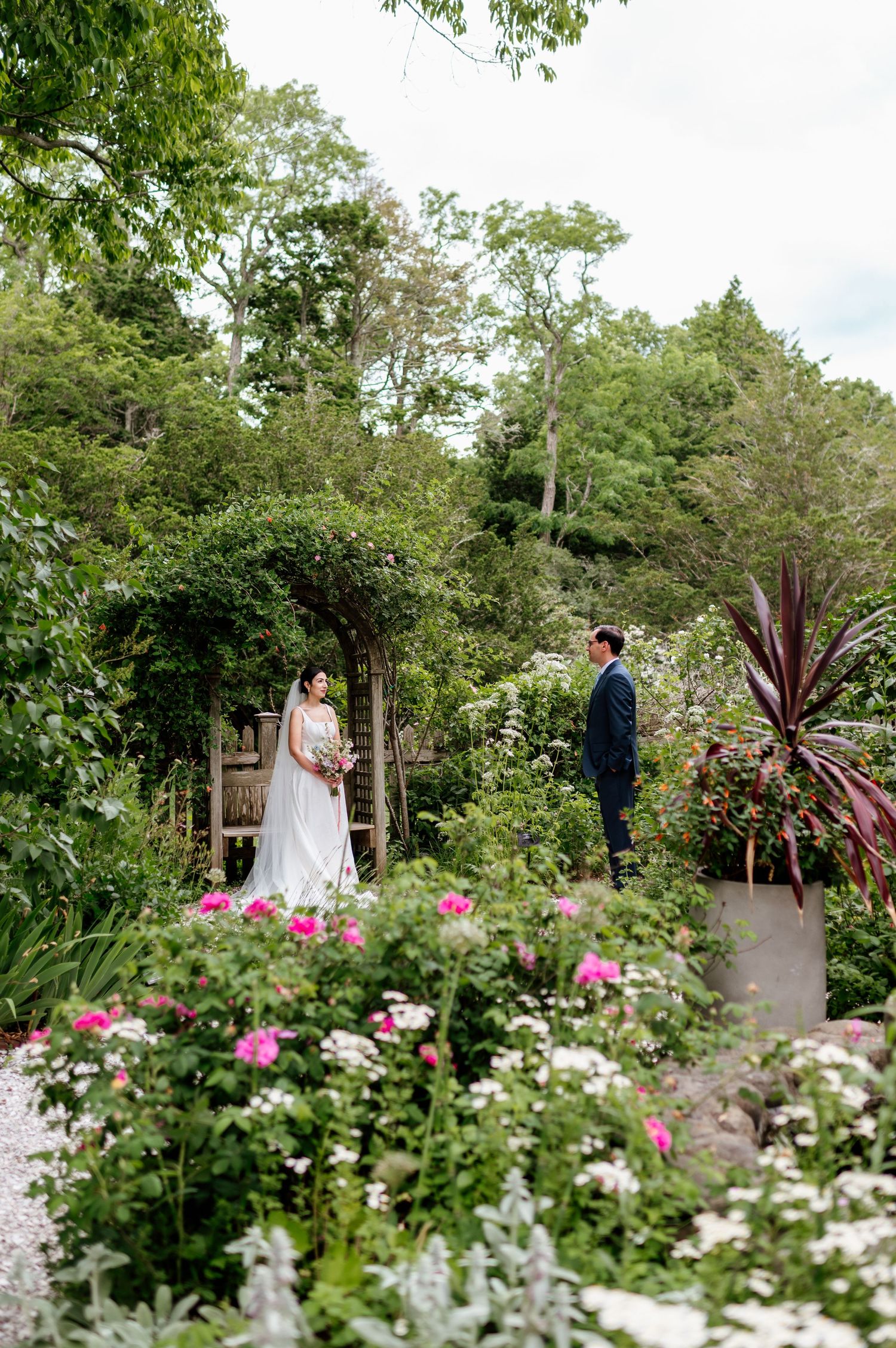 Bride and groom stand among lush garden blooms with pink roses and greenery at an outdoor wedding ceremony.
