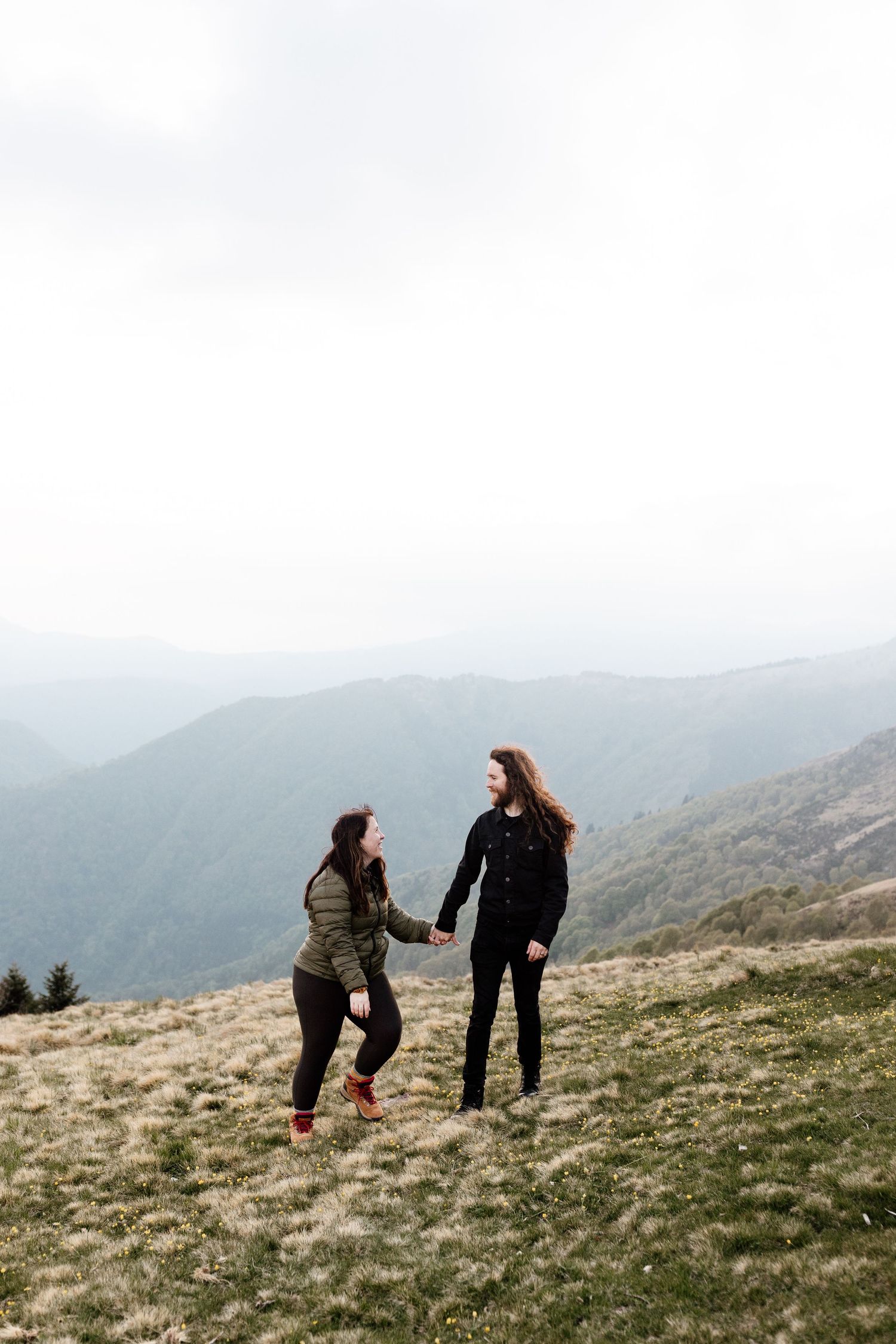 Two people bend down to examine something on a grassy mountainside with misty peaks in the background.