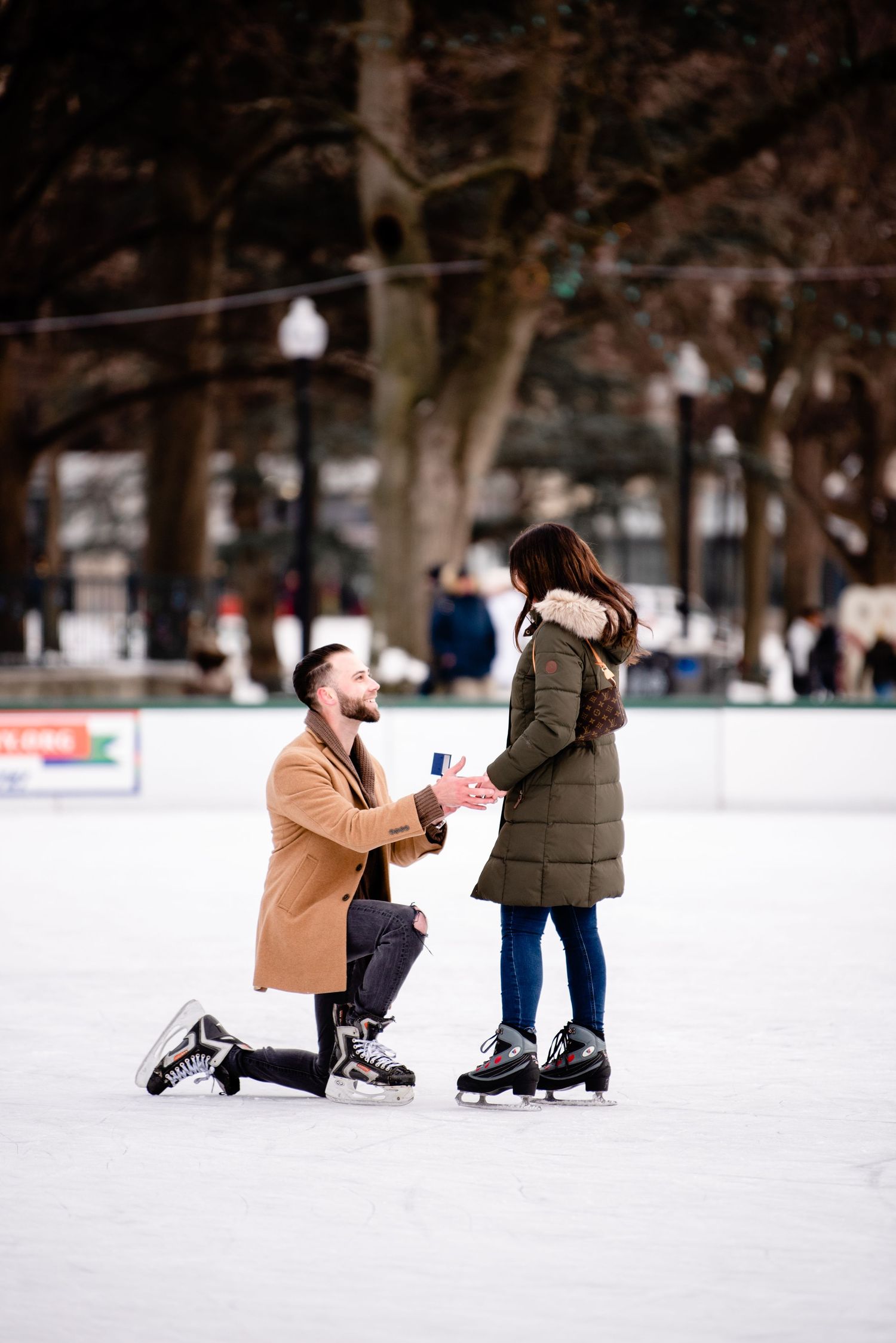 A winter proposal scene on an ice skating rink surrounded by snow-covered trees.