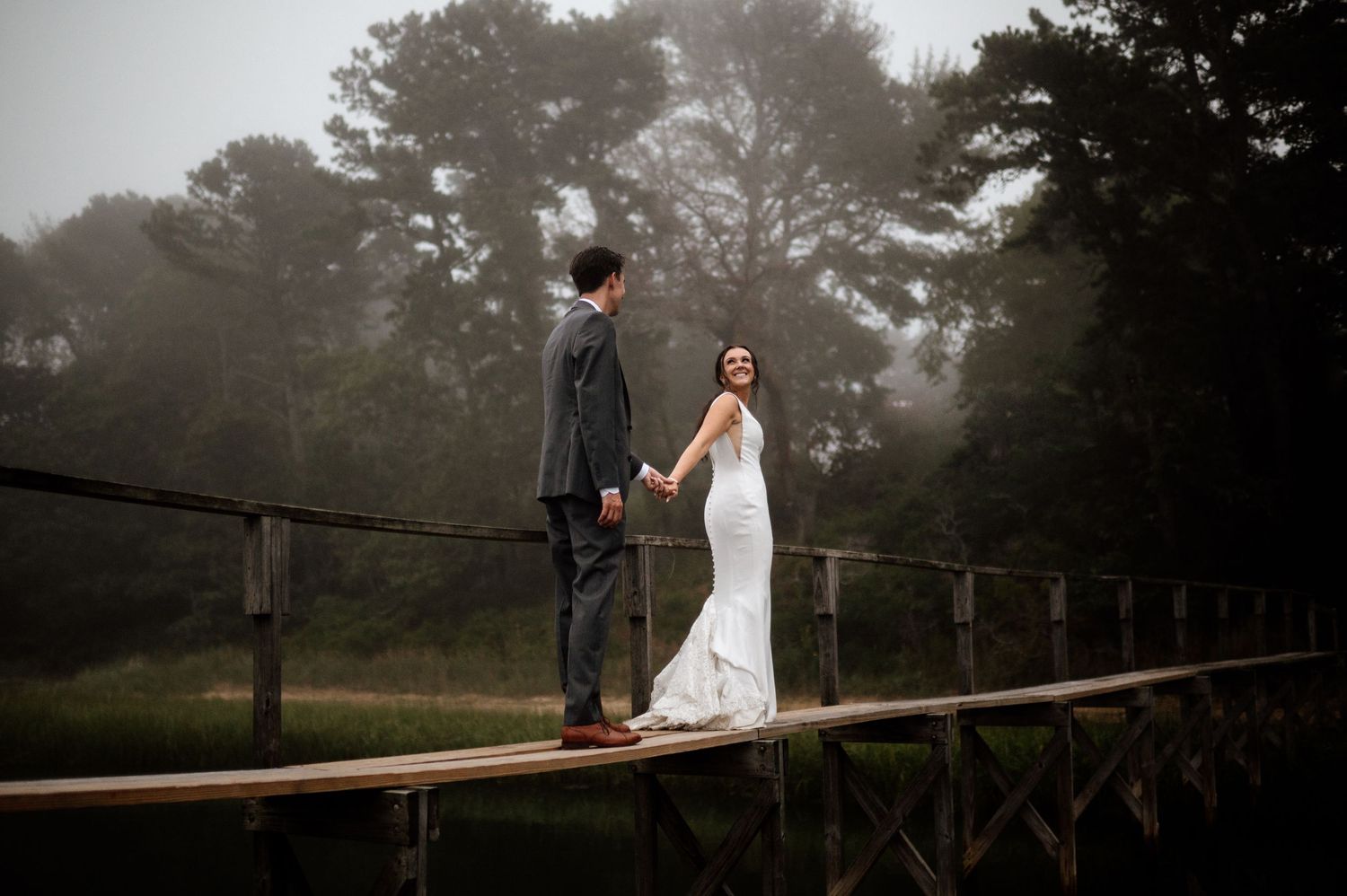 Wedding couple poses on wooden walkway in misty forest setting.