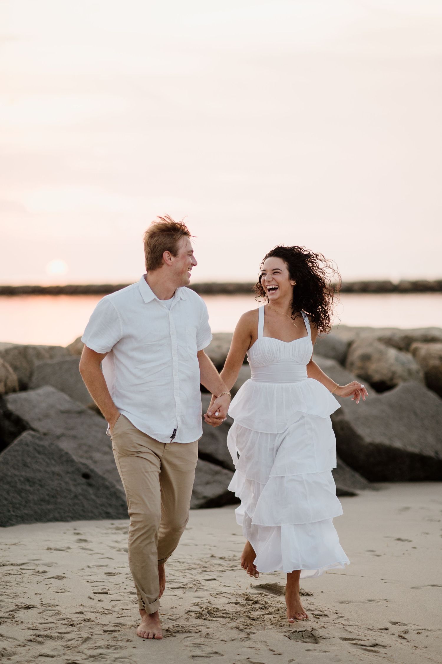 A joyful couple runs barefoot along a beach with rocks at sunset wearing white and beige outfits.