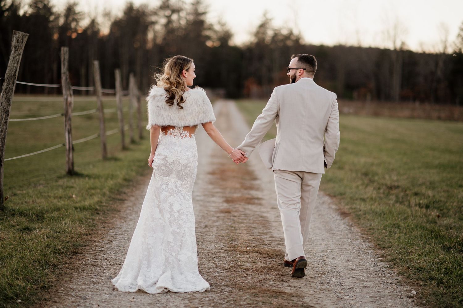A couple in wedding attire walks hand in hand down a rustic path during sunset.