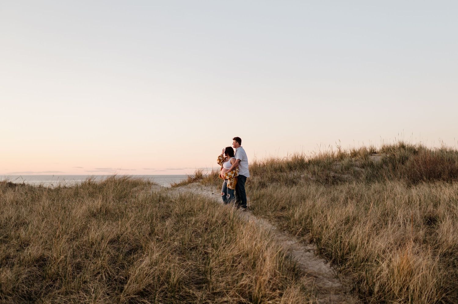 A couple stands together on a grassy hilltop path during golden hour sunset with tall dry grass surrounding them.