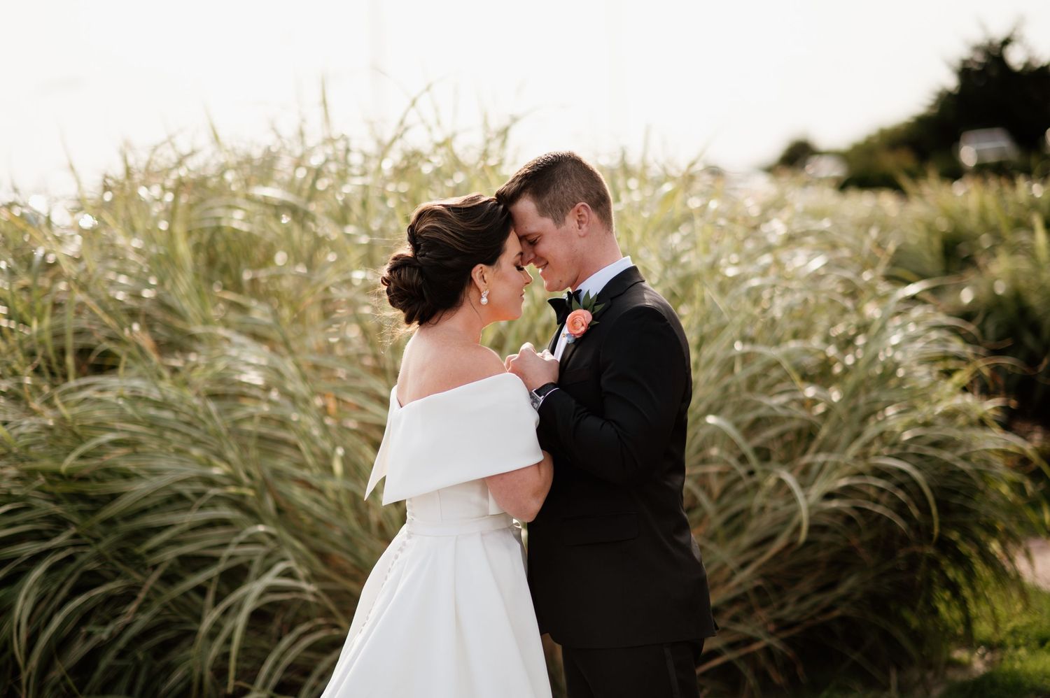 A romantic wedding couple shares an intimate moment among tall ornamental grasses at sunset.