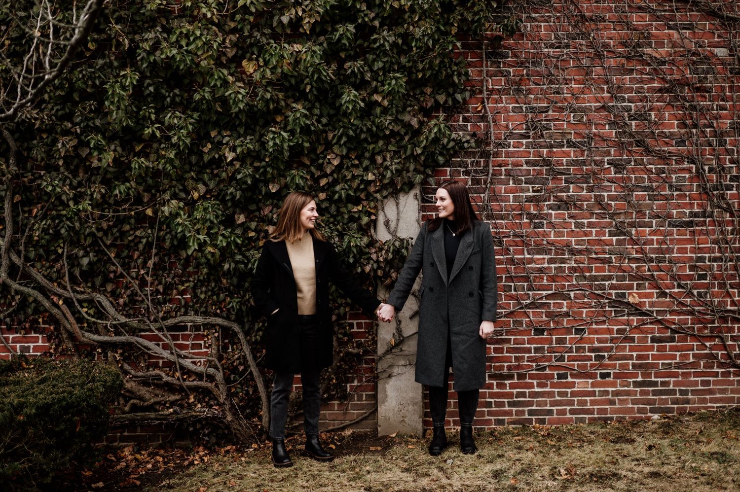 A couple shares a laugh while standing against a rustic brick wall surrounded by foliage.