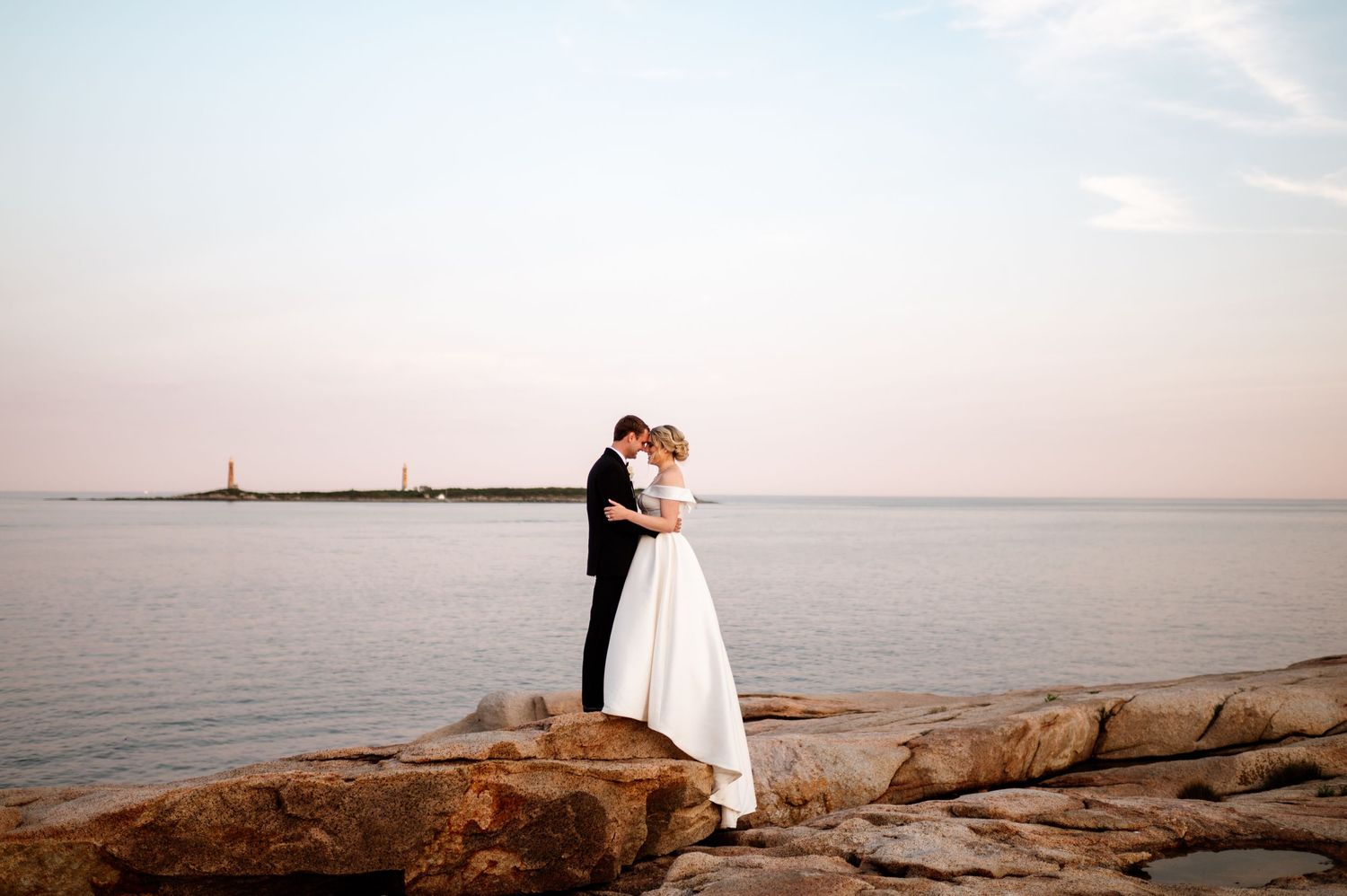 A romantic seaside wedding photoshoot with a couple standing on rocky coastal cliffs at sunset.