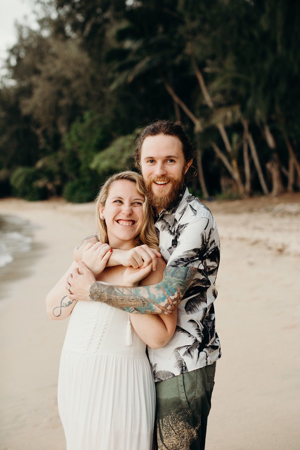 A couple embraces lovingly on a sandy beach with trees in the background.