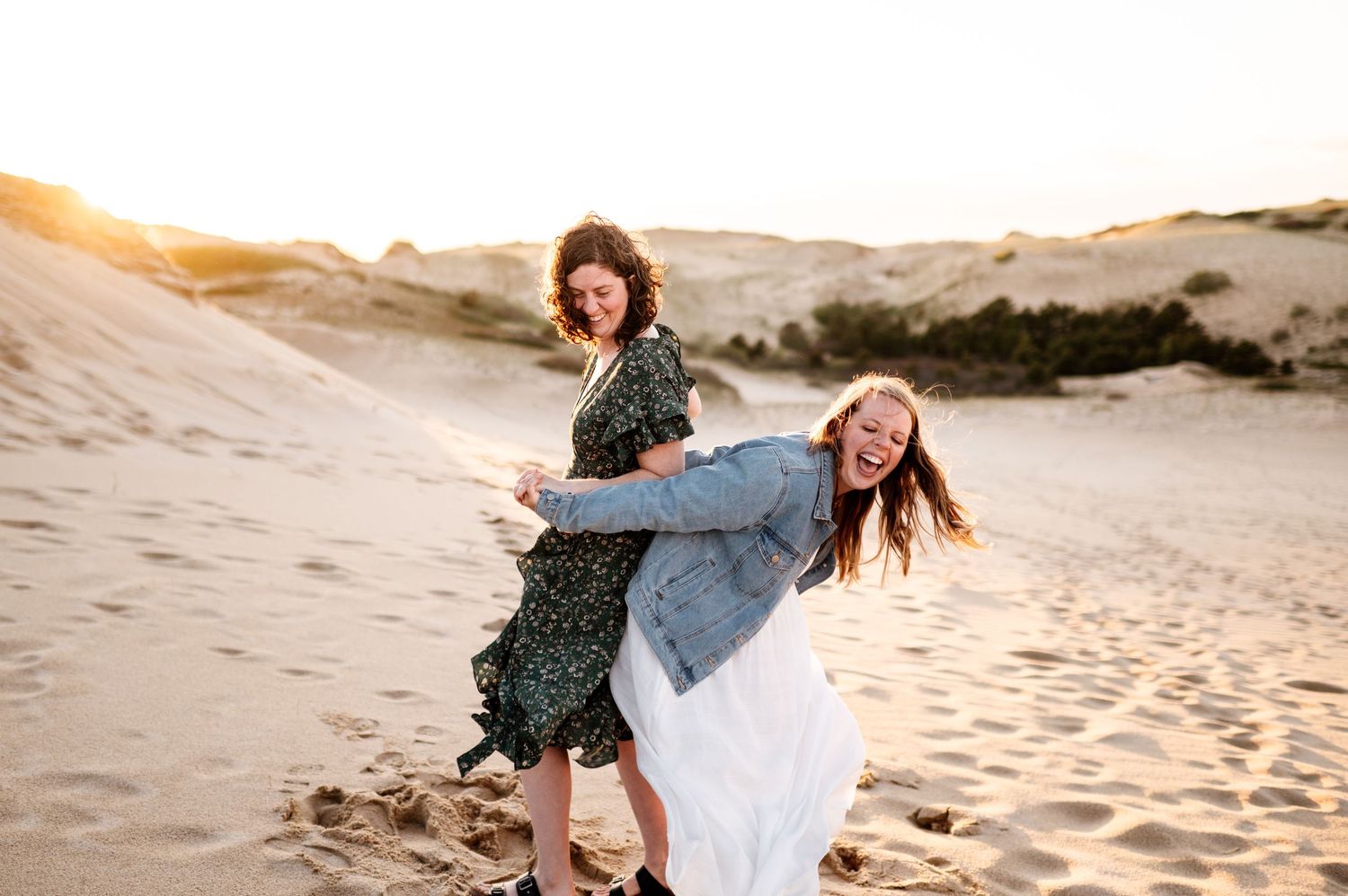 Two friends playing and laughing together on sandy dunes at sunset.