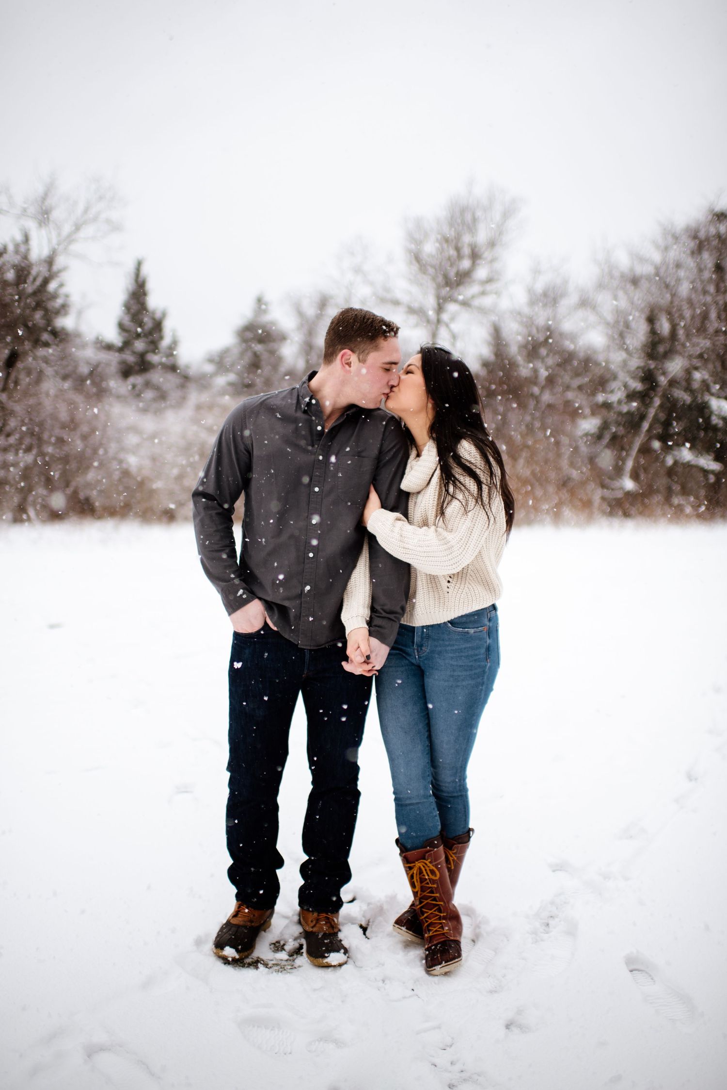Couple shares sweet winter moment in snowy field with snowflakes falling around them.