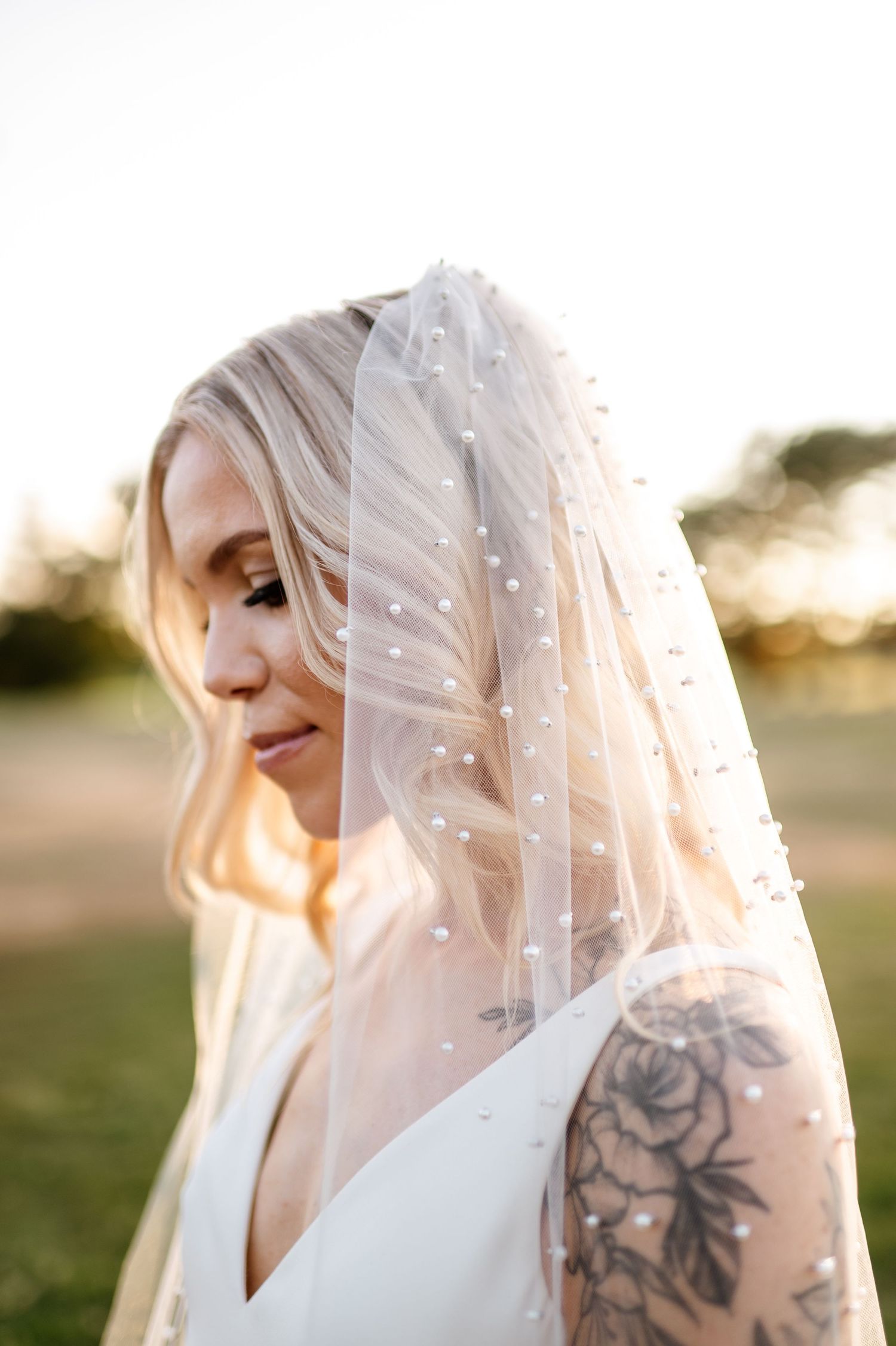 A bride in a white dress and delicate pearl-dotted veil poses in soft golden sunlight, showing a floral arm tattoo.