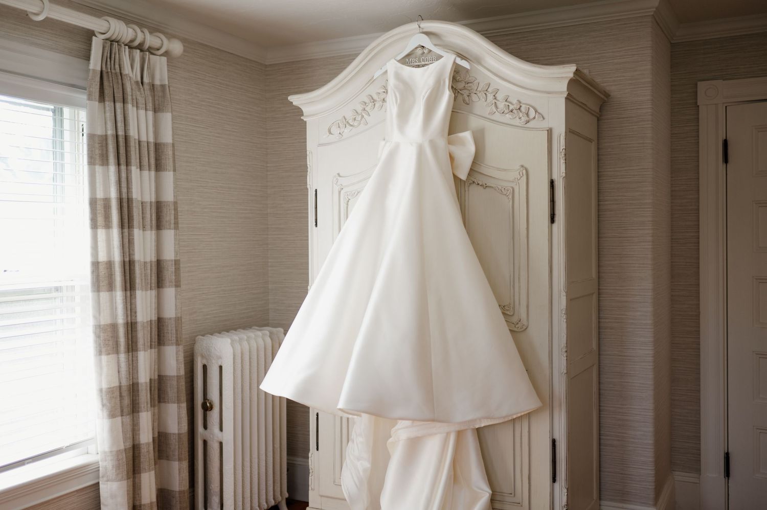 A vintage white christening gown hangs elegantly near a window, showcasing delicate lace details and traditional craftsmanship.