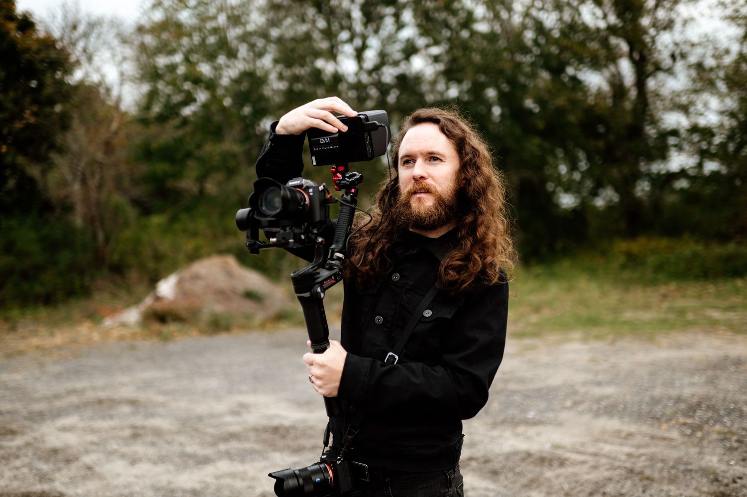 A photographer with long hair holds a professional camera outdoors in a natural setting.