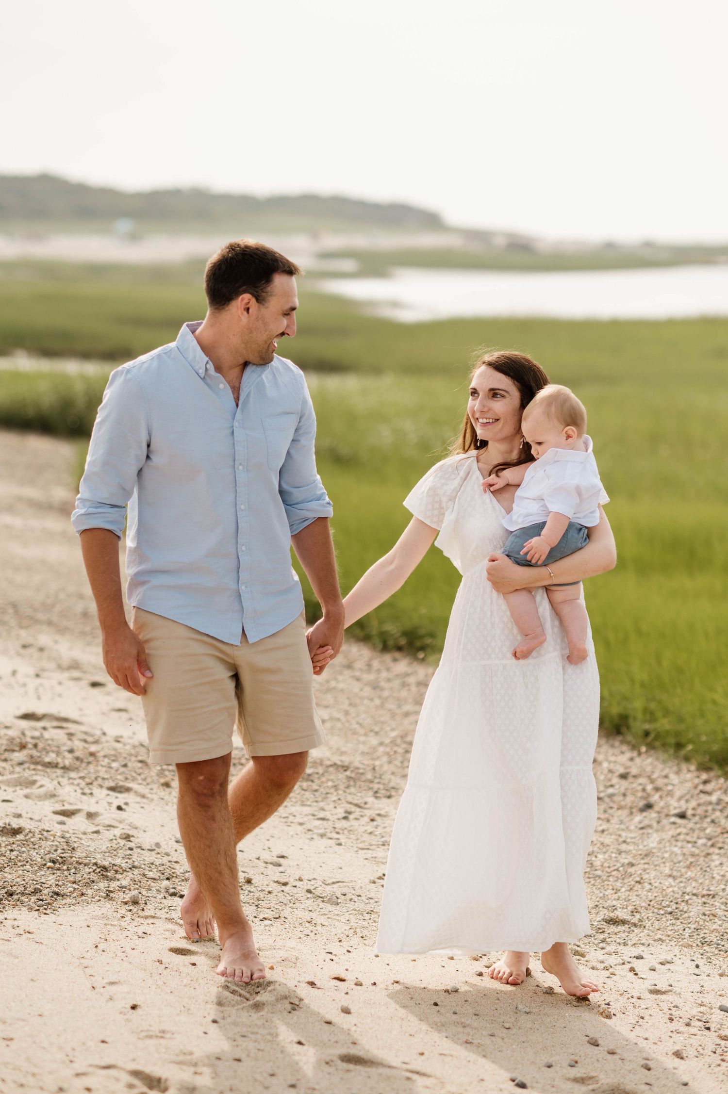 A family walks barefoot together on a sandy beach path surrounded by green coastal marshland on a summer day.