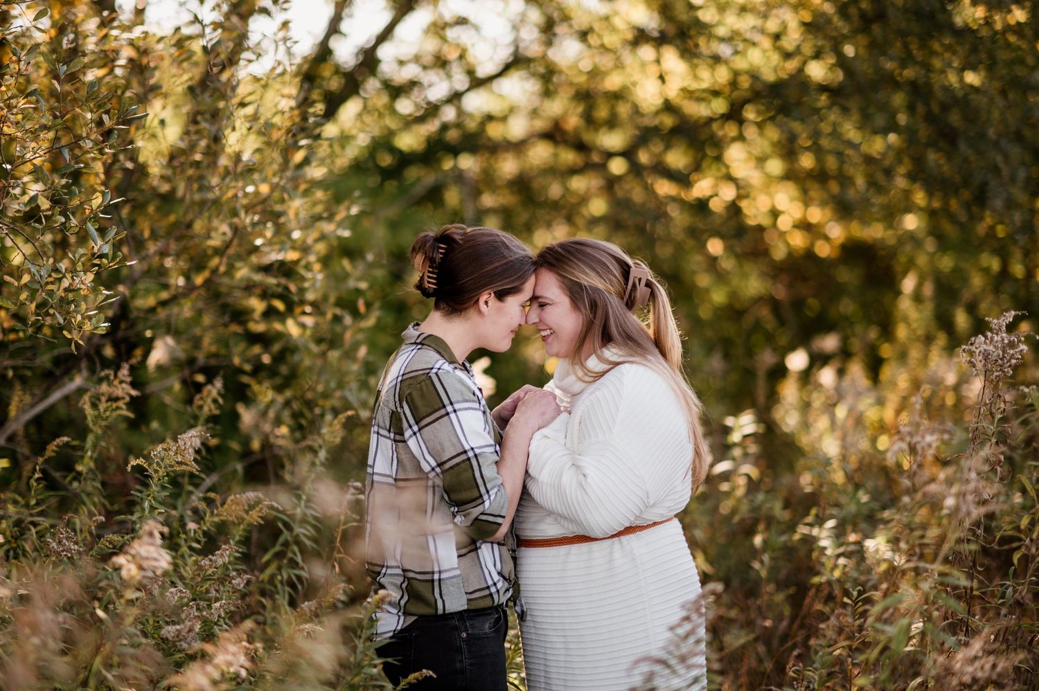 Romantic couple embraces in autumn field with golden sunlight filtering through trees during engagement session.
