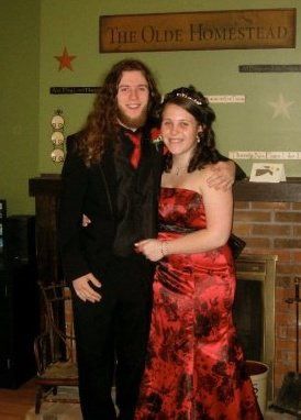 A couple poses together at The Olde Homestead, with one wearing a striking red dress and the other in dark attire.