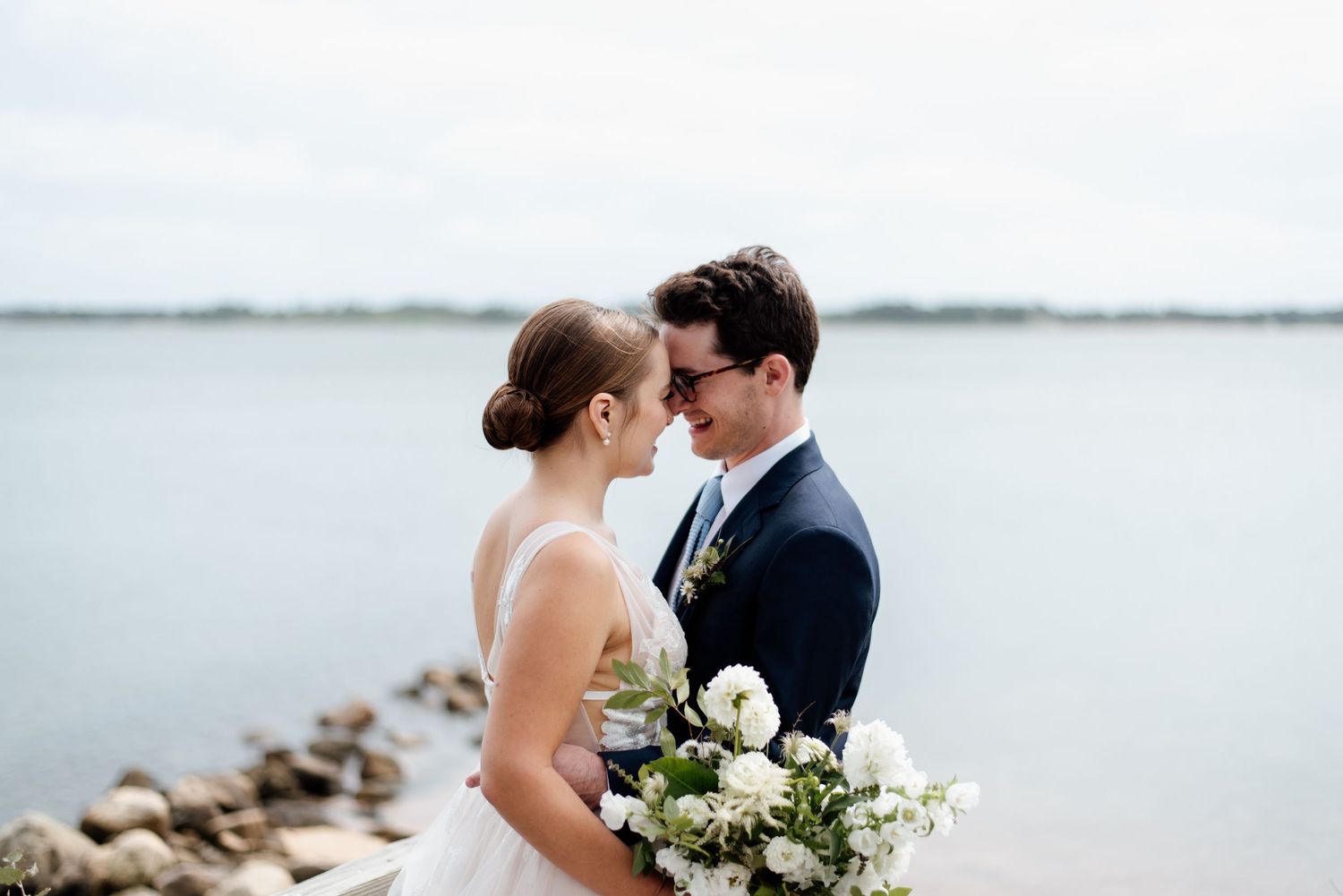 A couple in wedding attire share an intimate moment by a rocky shoreline with white flowers and a serene lake backdrop.