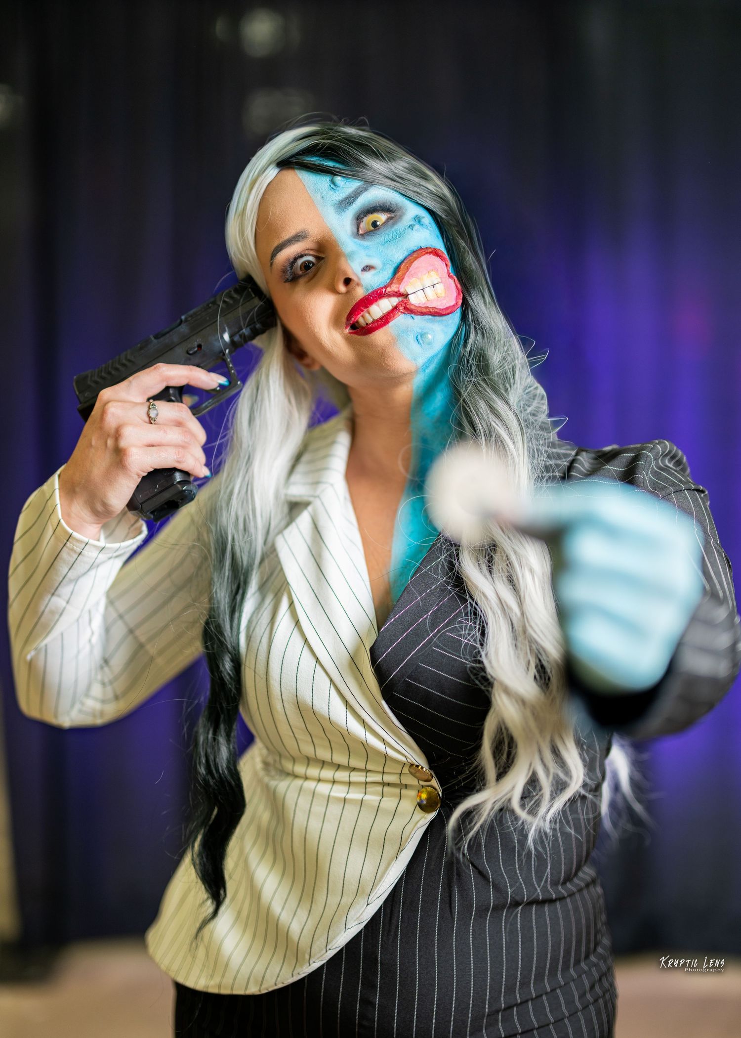 Cosplayer in pinstriped suit jacket with two-toned hair poses dramatically against purple backdrop.