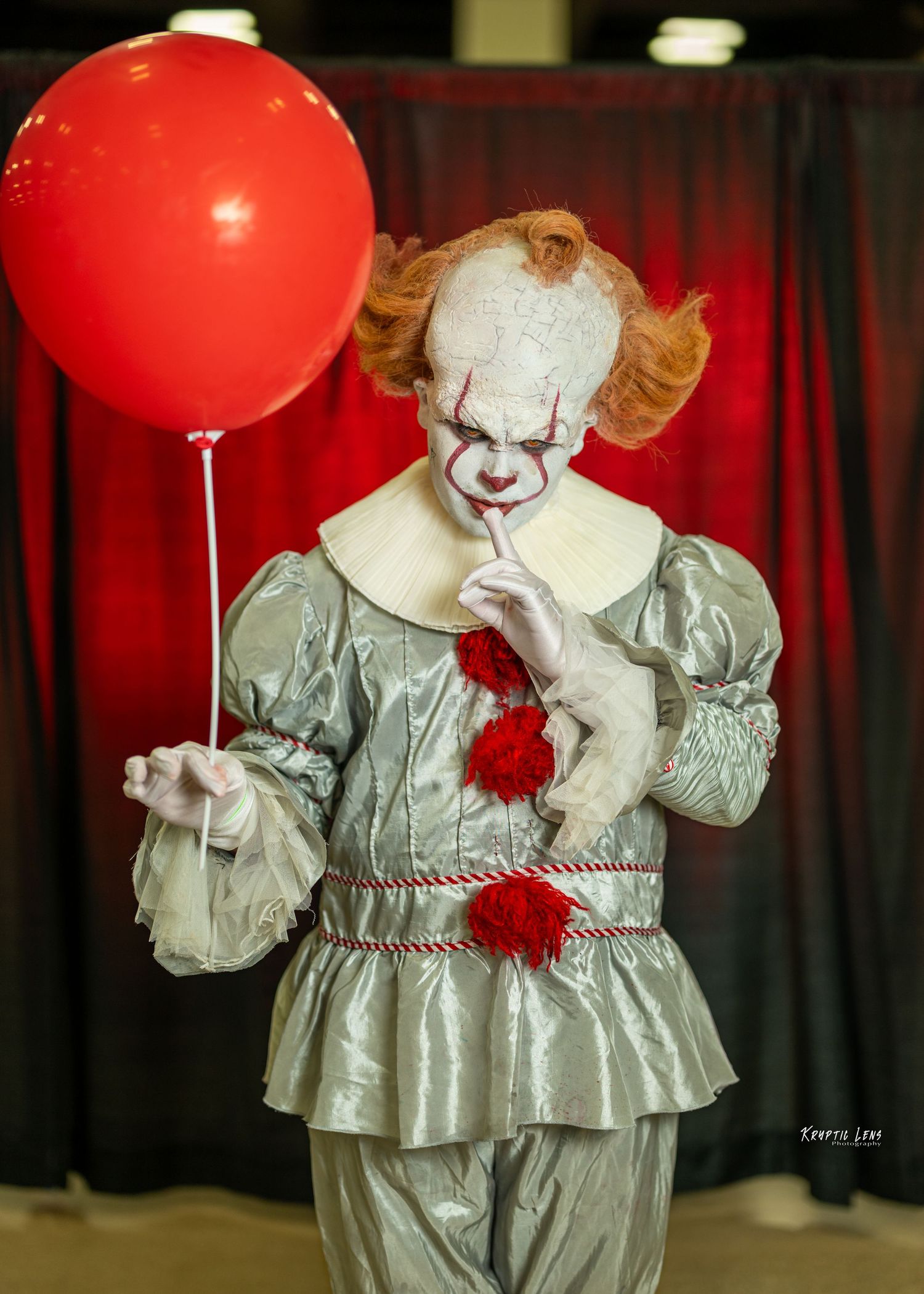 Creepy clown costume with silver outfit holding red balloon against dark curtain background.