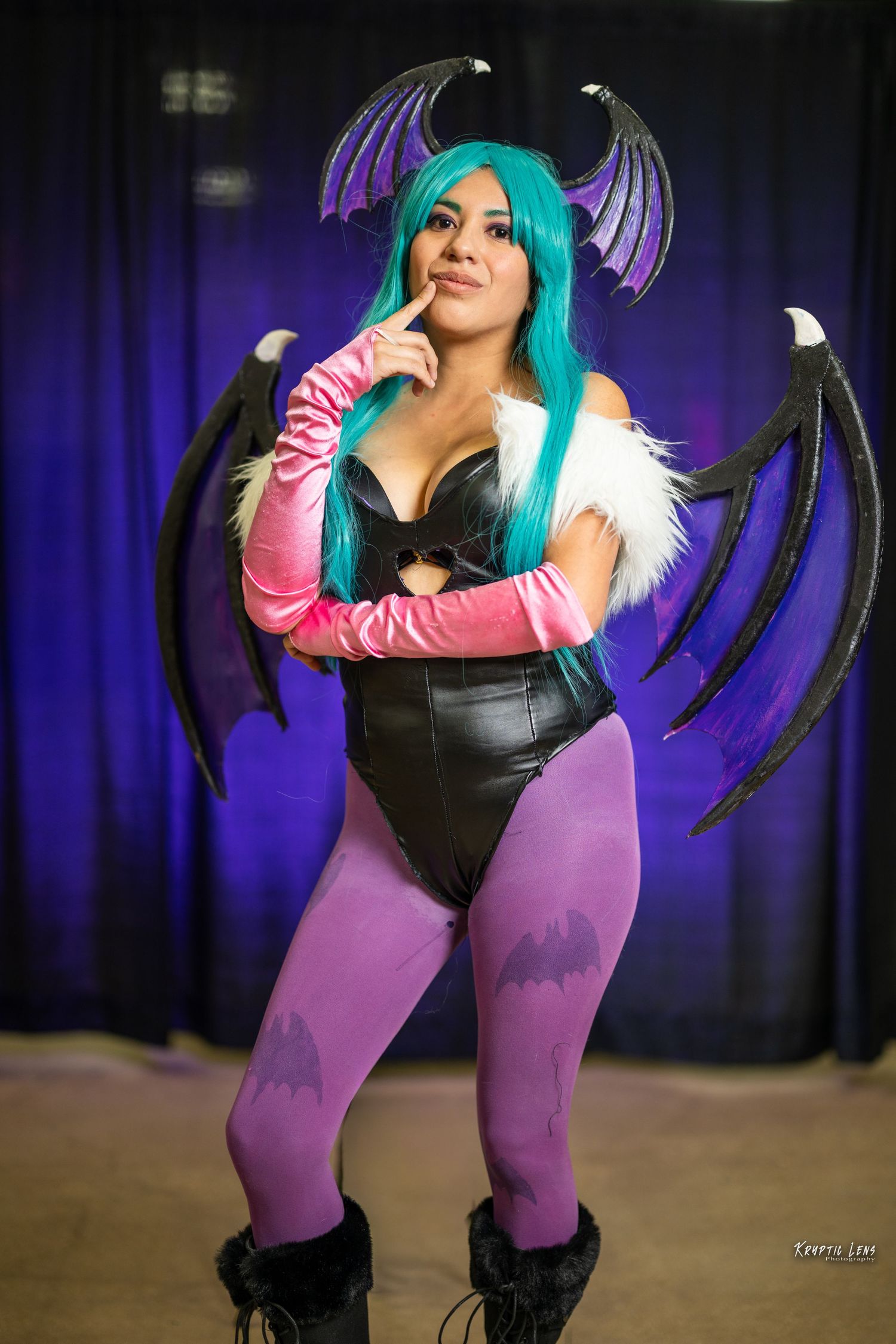Cosplayer in purple tights and black corset poses with teal wig and dark wings against blue backdrop.