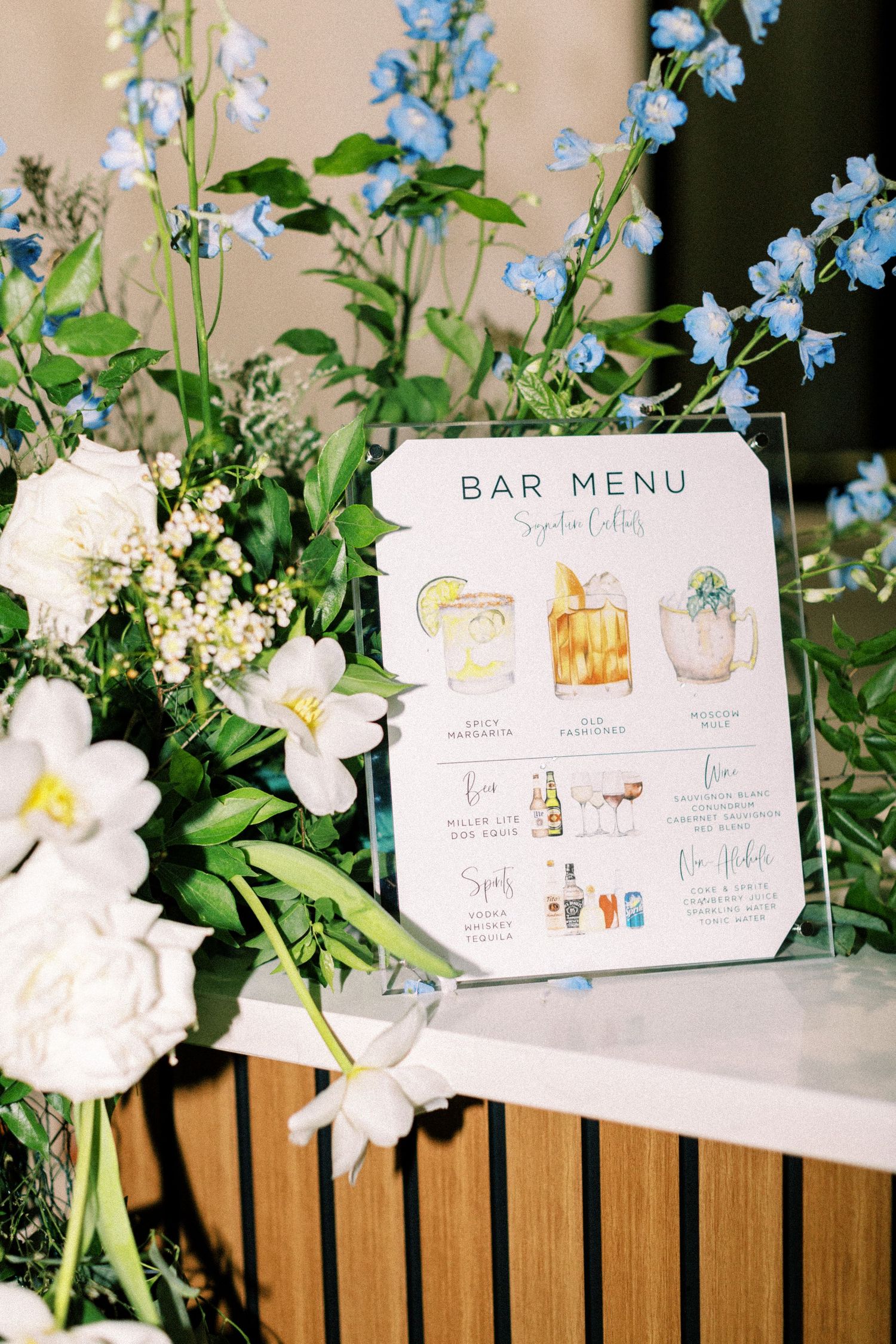 Elegant wedding bar menu with watercolor illustrations displayed among white and blue flowers.