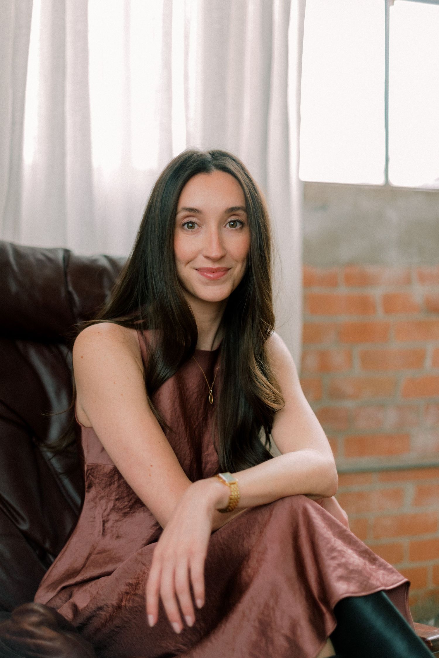 A person in a brown satin dress sits casually on a dark leather couch near exposed brick walls and white curtains.