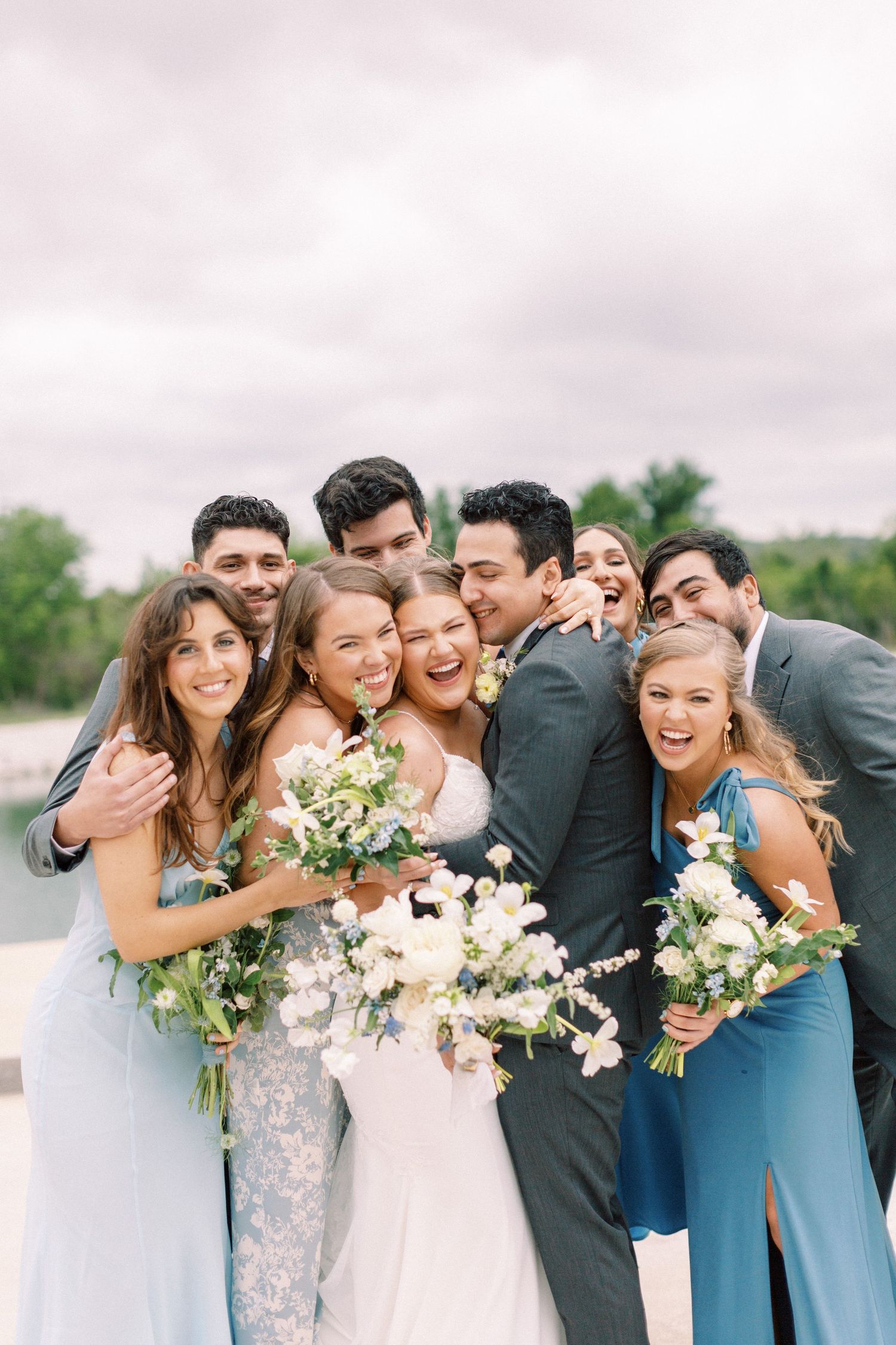 Wedding party in blue dresses and gray suits posing together with white bouquets outdoors.