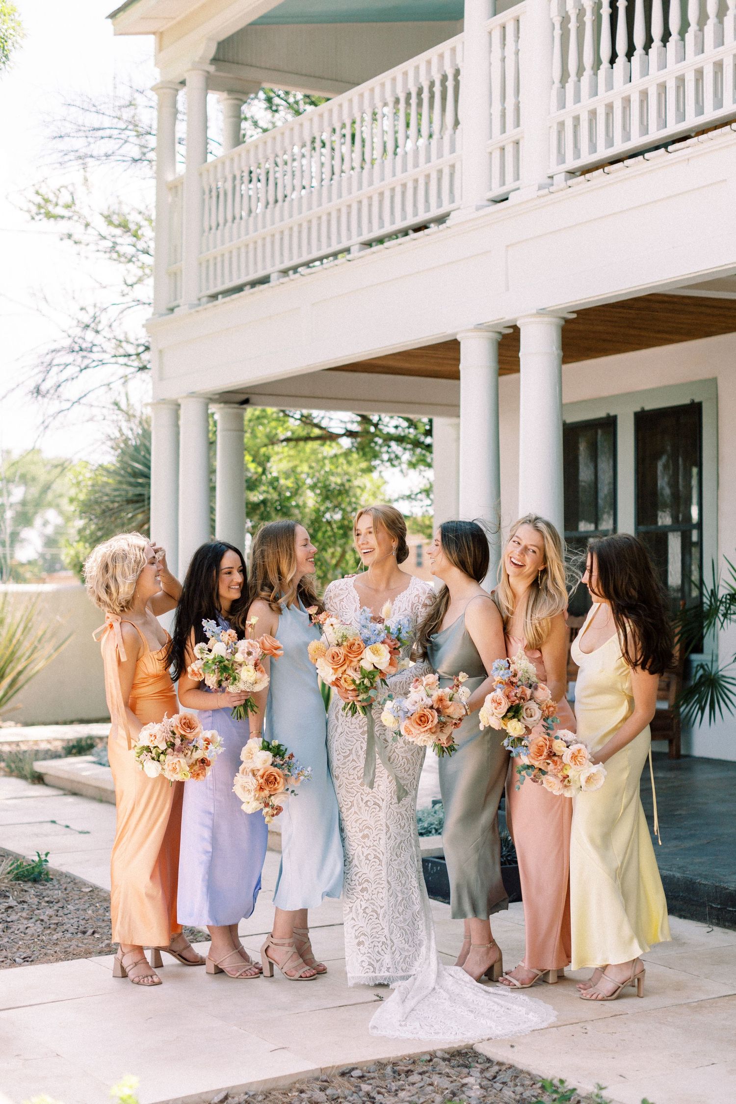 Bridesmaids in pastel dresses lined up on a white columned porch holding colorful bouquets.