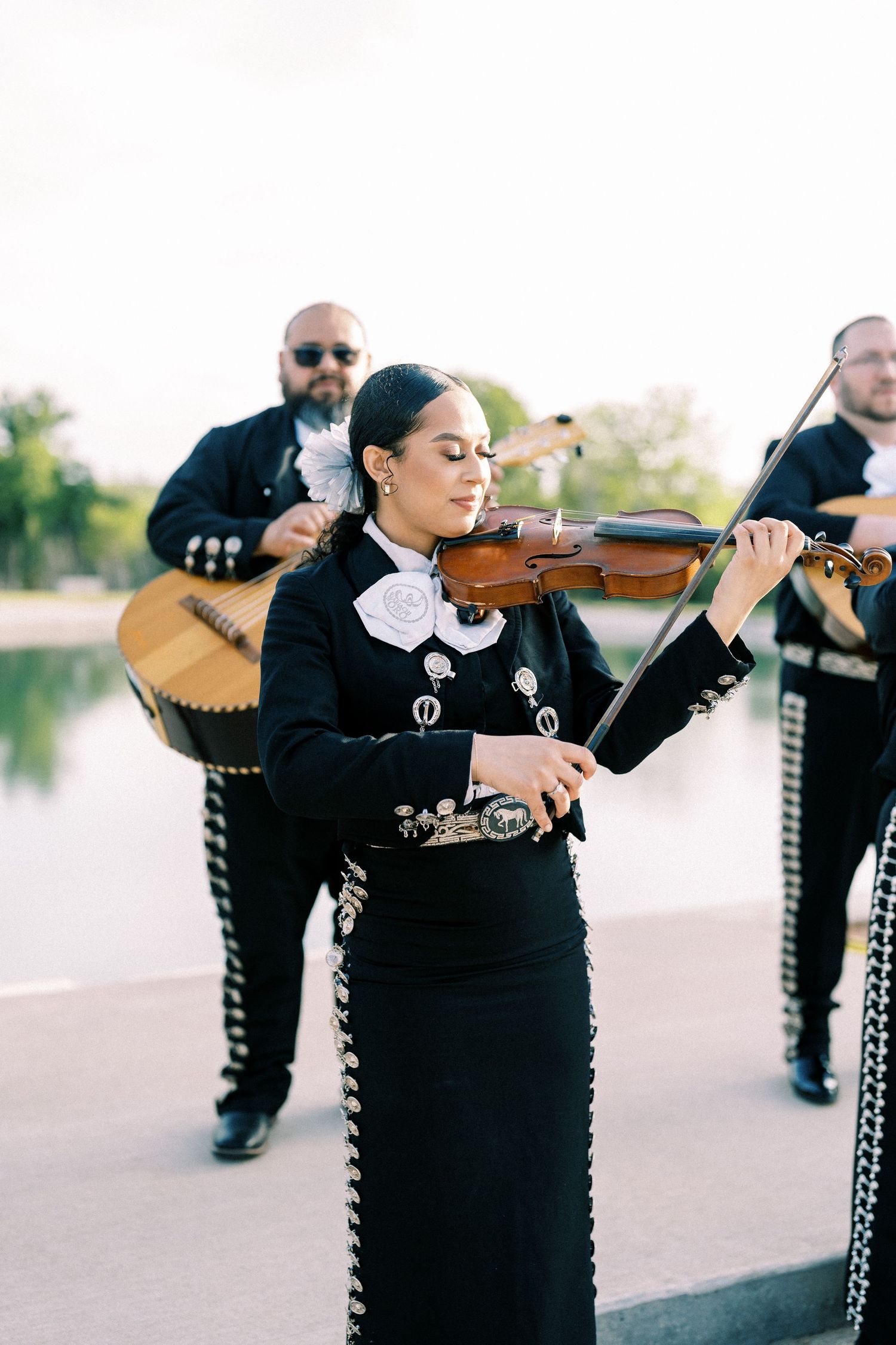 Mariachi band members in traditional black outfits playing violins outdoors.
