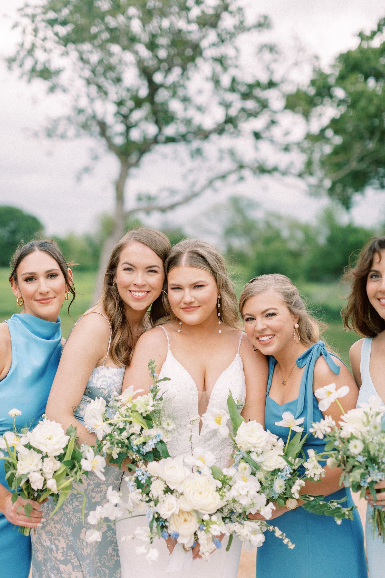 Bridesmaids in light blue dresses hold elegant white and green bouquets while posing outdoors.