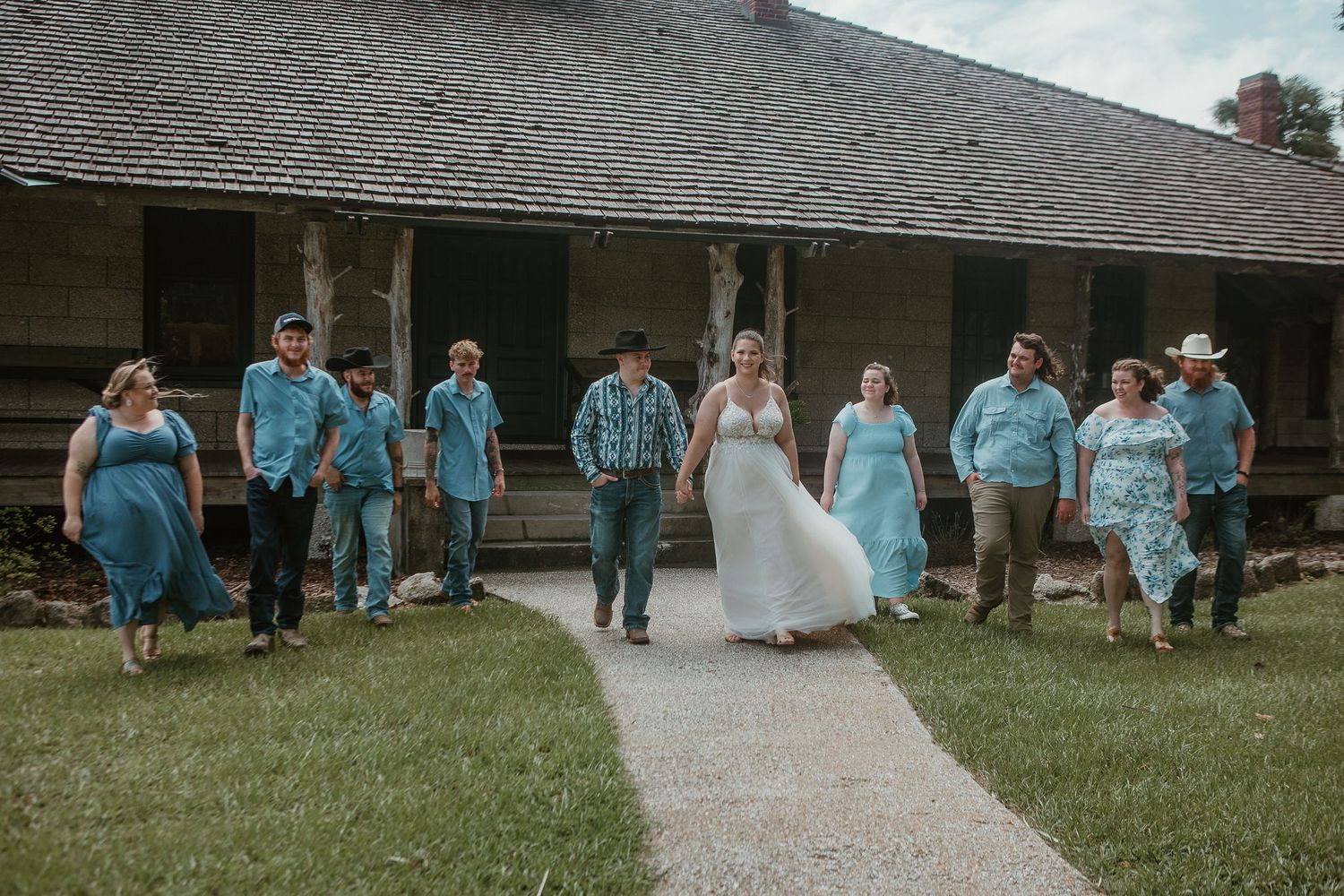 Wedding party members in light blue dresses walking along a path outside a building.