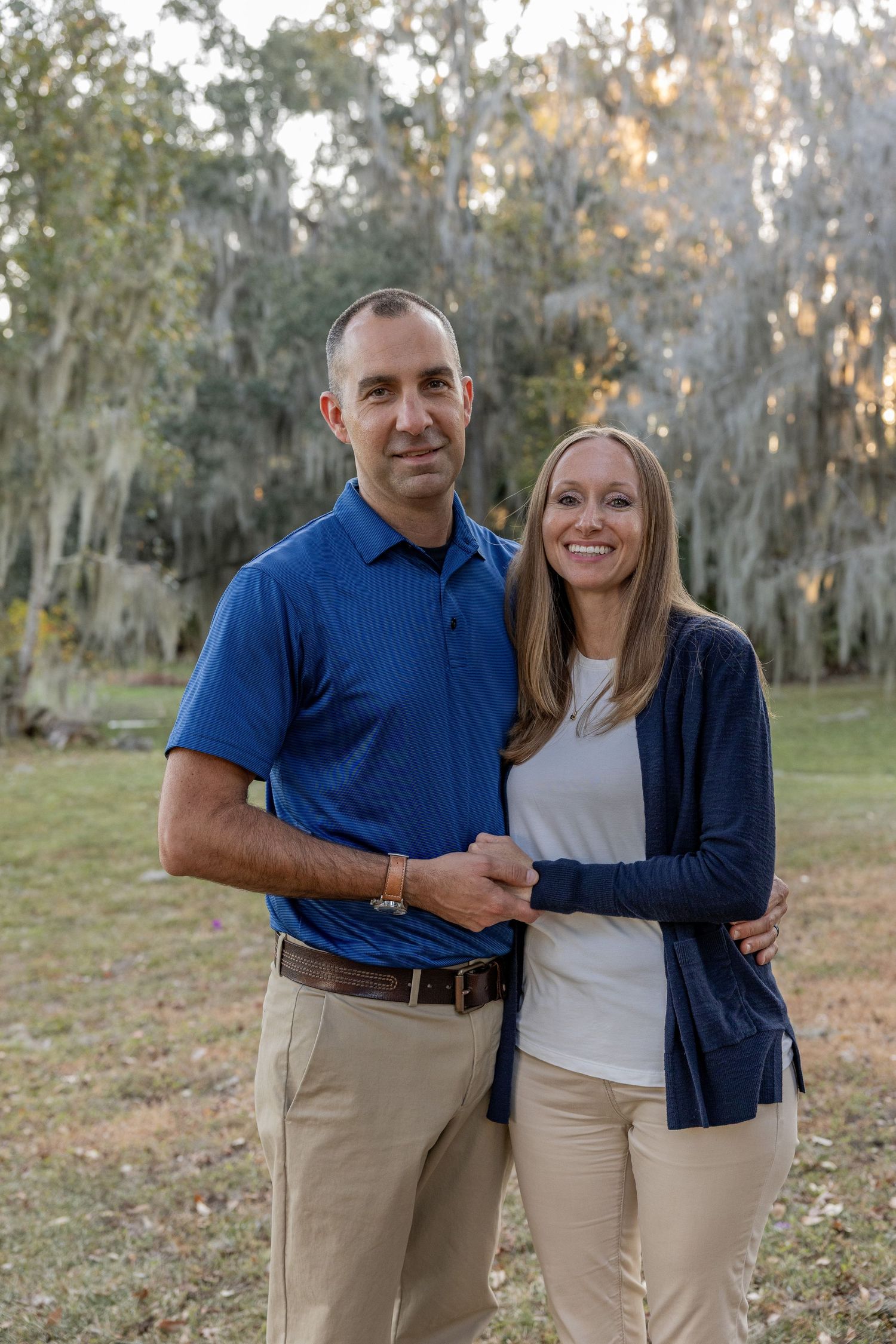 A couple poses together outdoors wearing coordinating blue and khaki outfits in a natural setting.