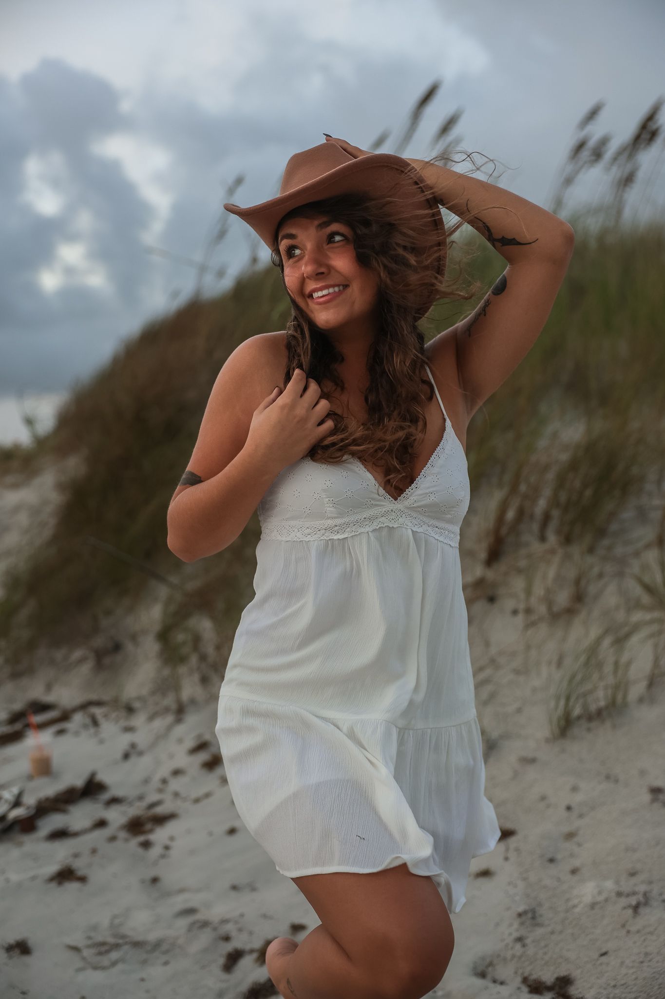 Woman in white dress poses playfully on sandy beach dunes during golden hour.