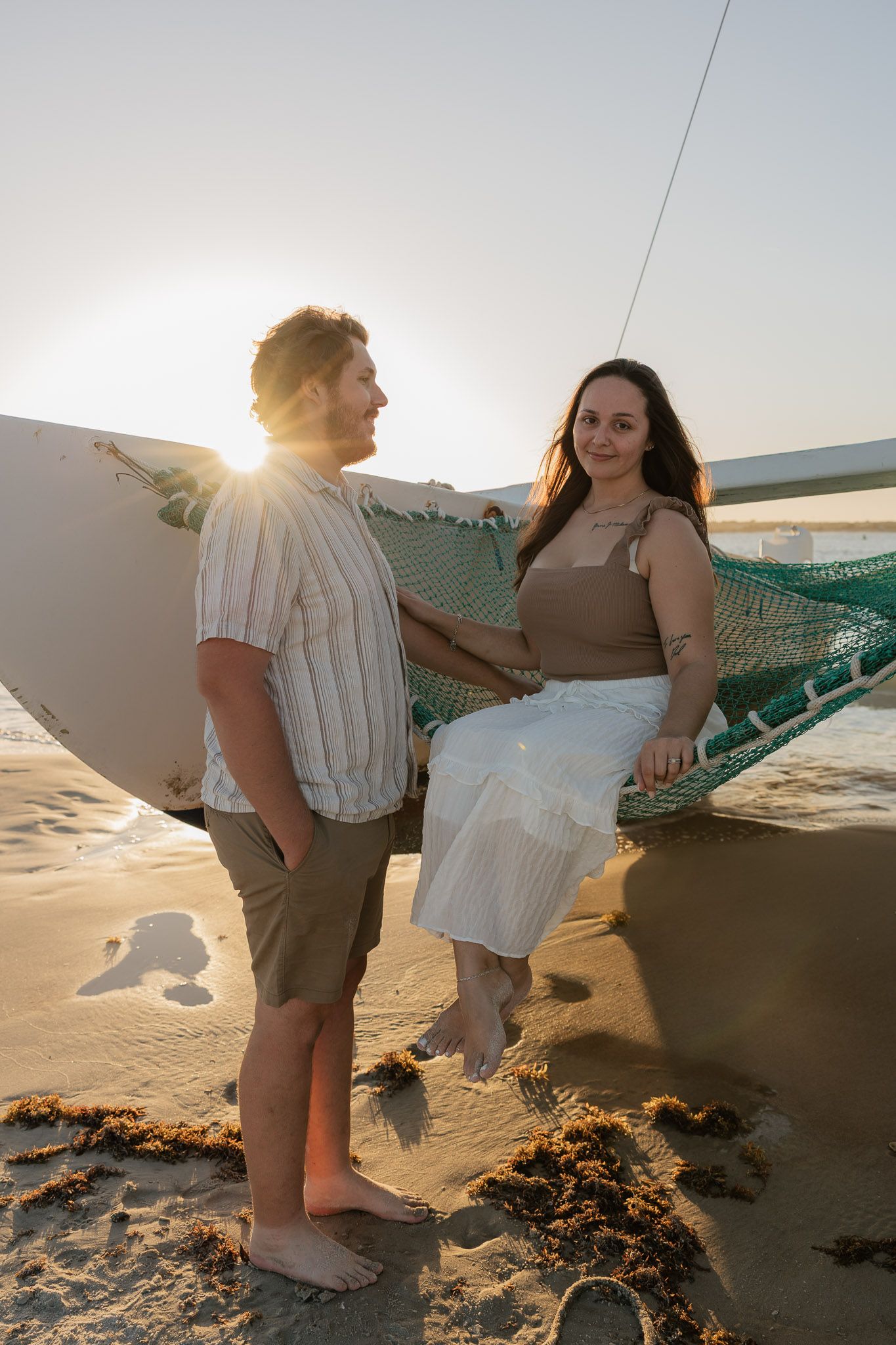 Couple on a sandy beach at sunset, woman sitting on hammock, man holding surfboard in golden light.
