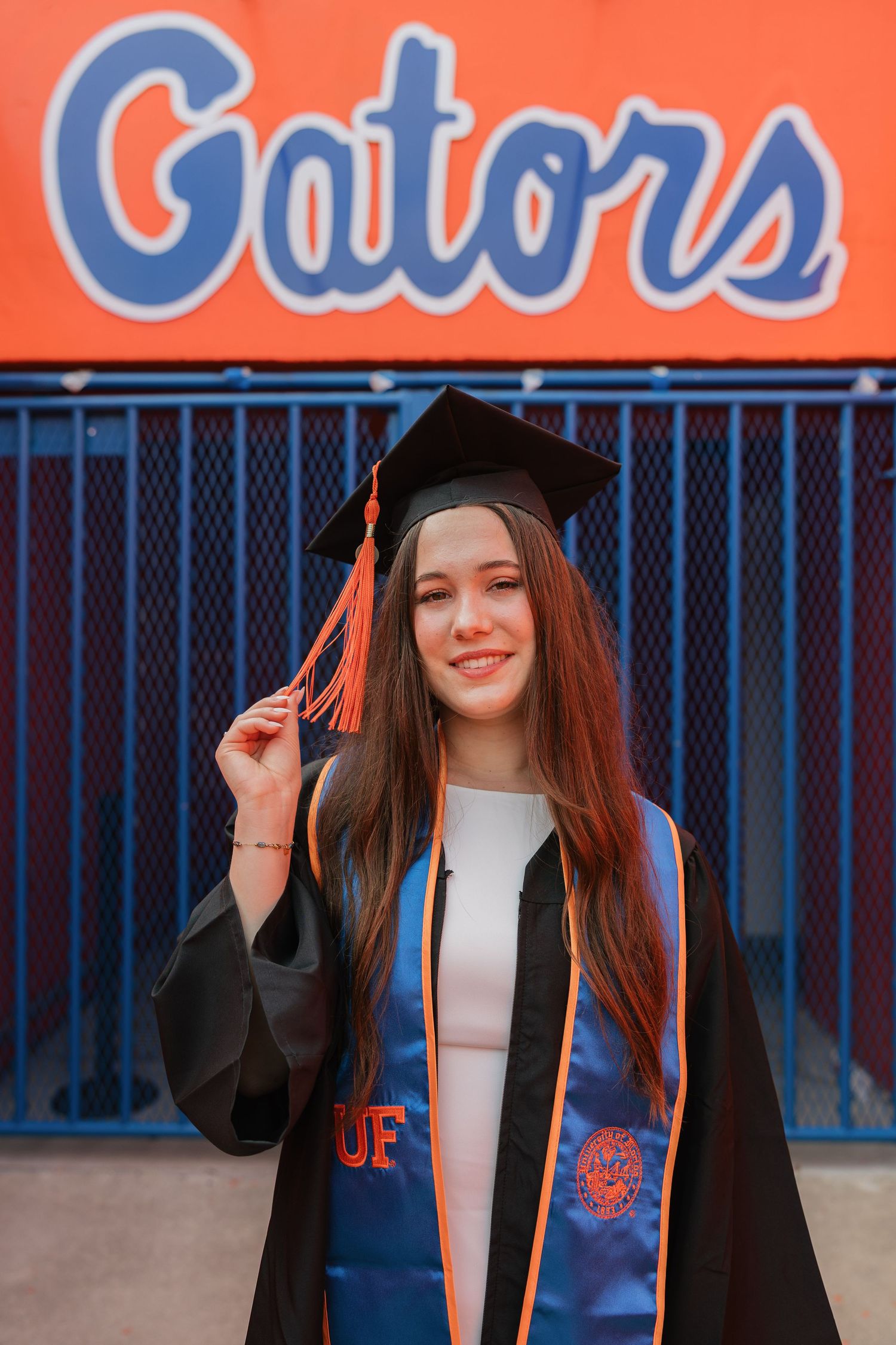 University of Florida graduate smiling in cap and gown with blue UF stole in front of orange Gators sign.