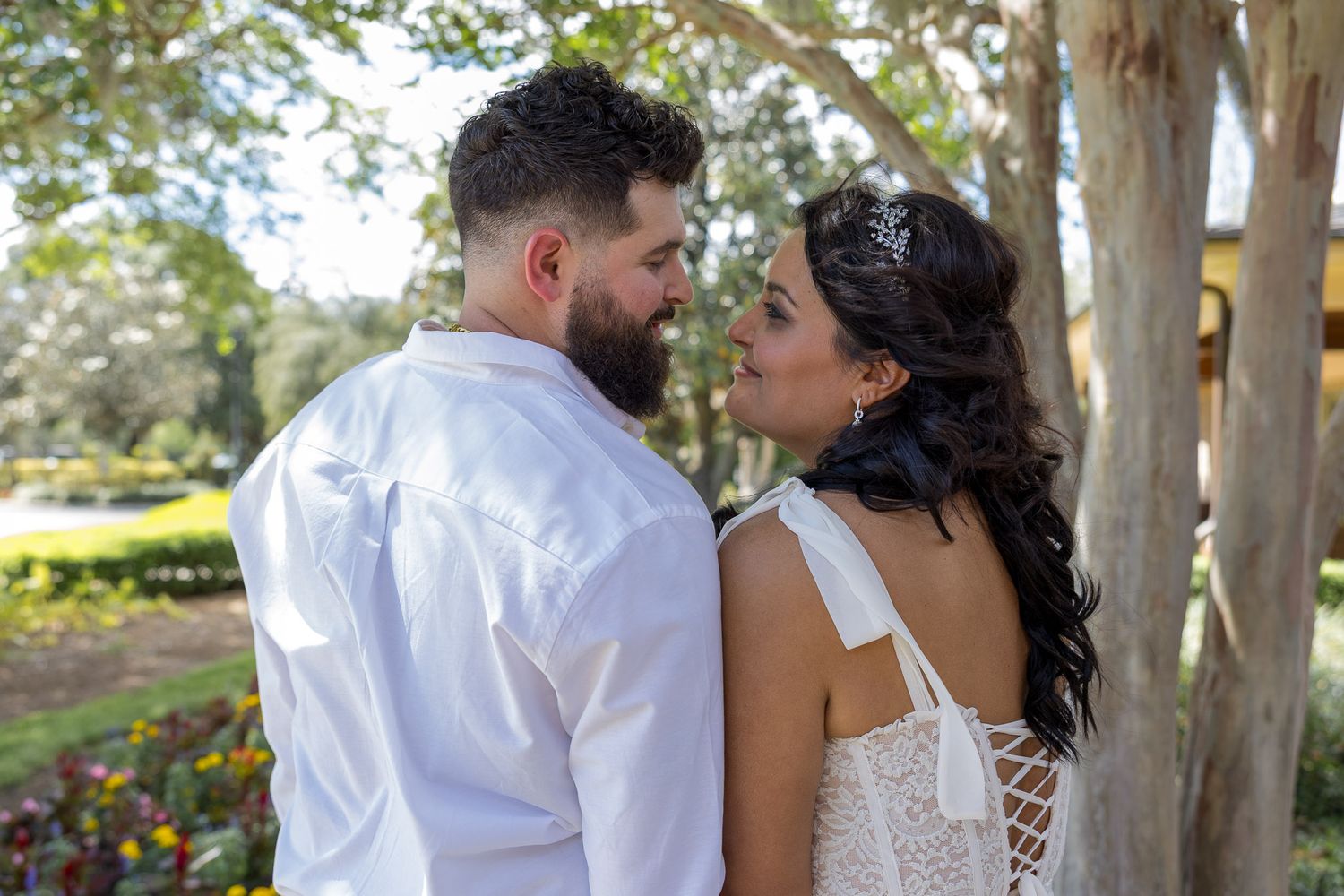 A couple in white wedding attire share a romantic moment outdoors among trees in a lush garden setting.