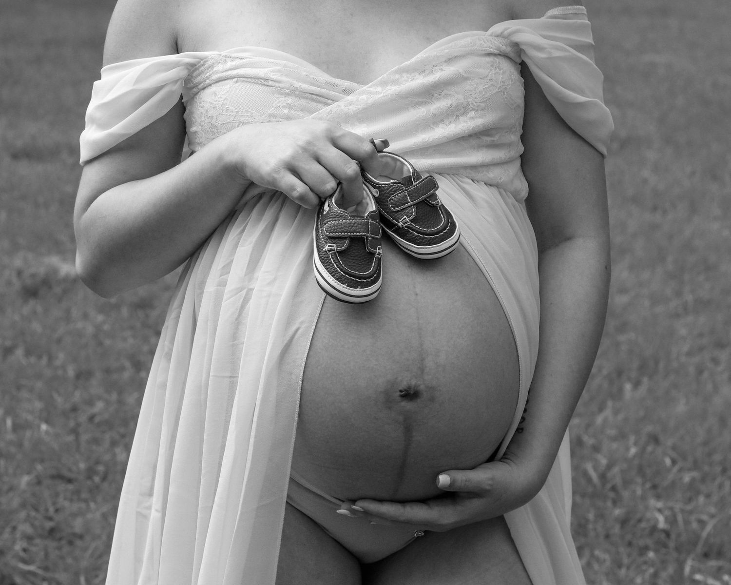 Black and white photo of pregnant woman in flowy dress holding baby shoes against her belly.