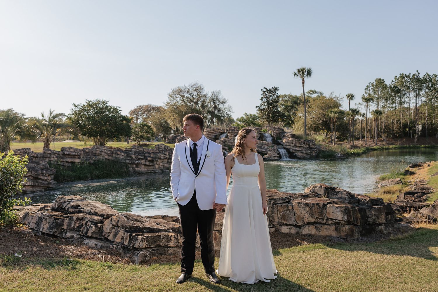 Bride and groom stand hand in hand by a scenic rocky pond with lush greenery at an outdoor wedding venue.
