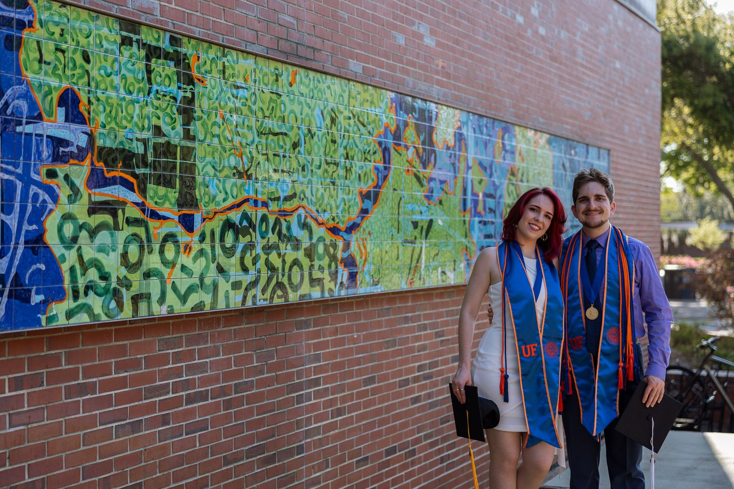 Two graduates in colorful stoles pose proudly in front of a vibrant mosaic mural on a brick wall.