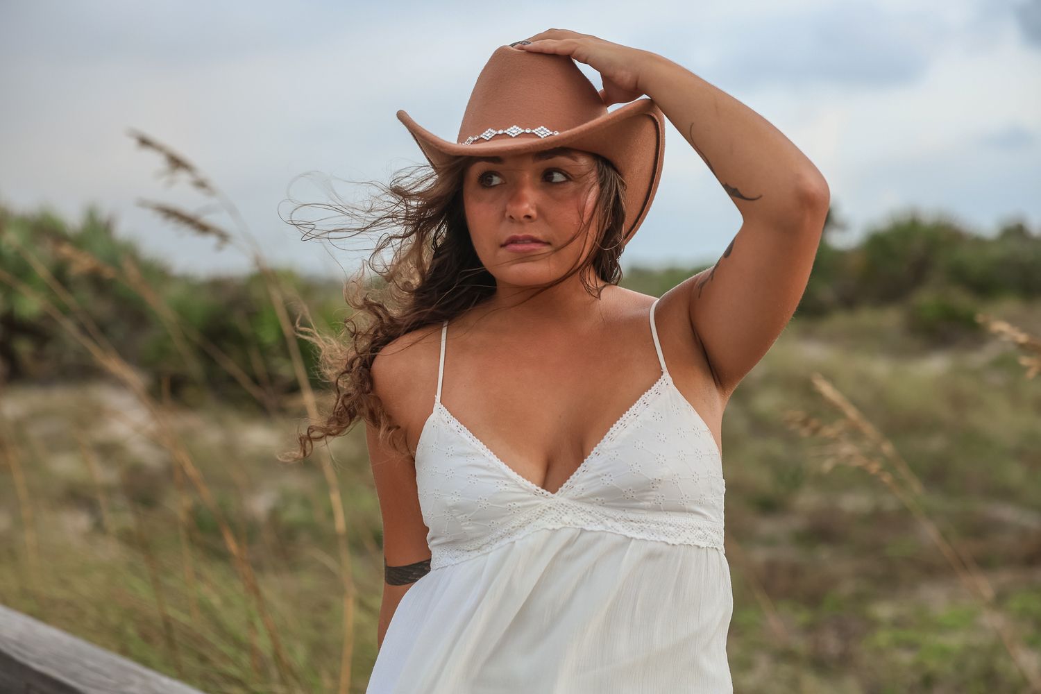A person in a white dress poses in tall grass against a cloudy sky.