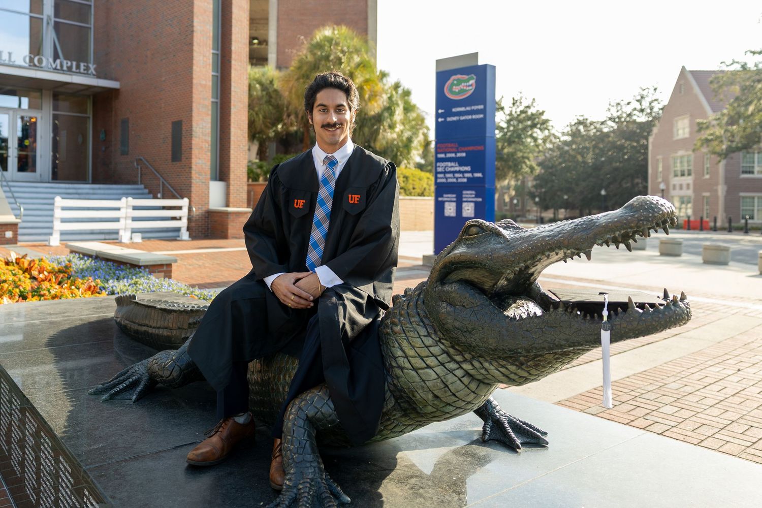 Graduate in formal attire poses on brick walkway surrounded by oak trees on campus.